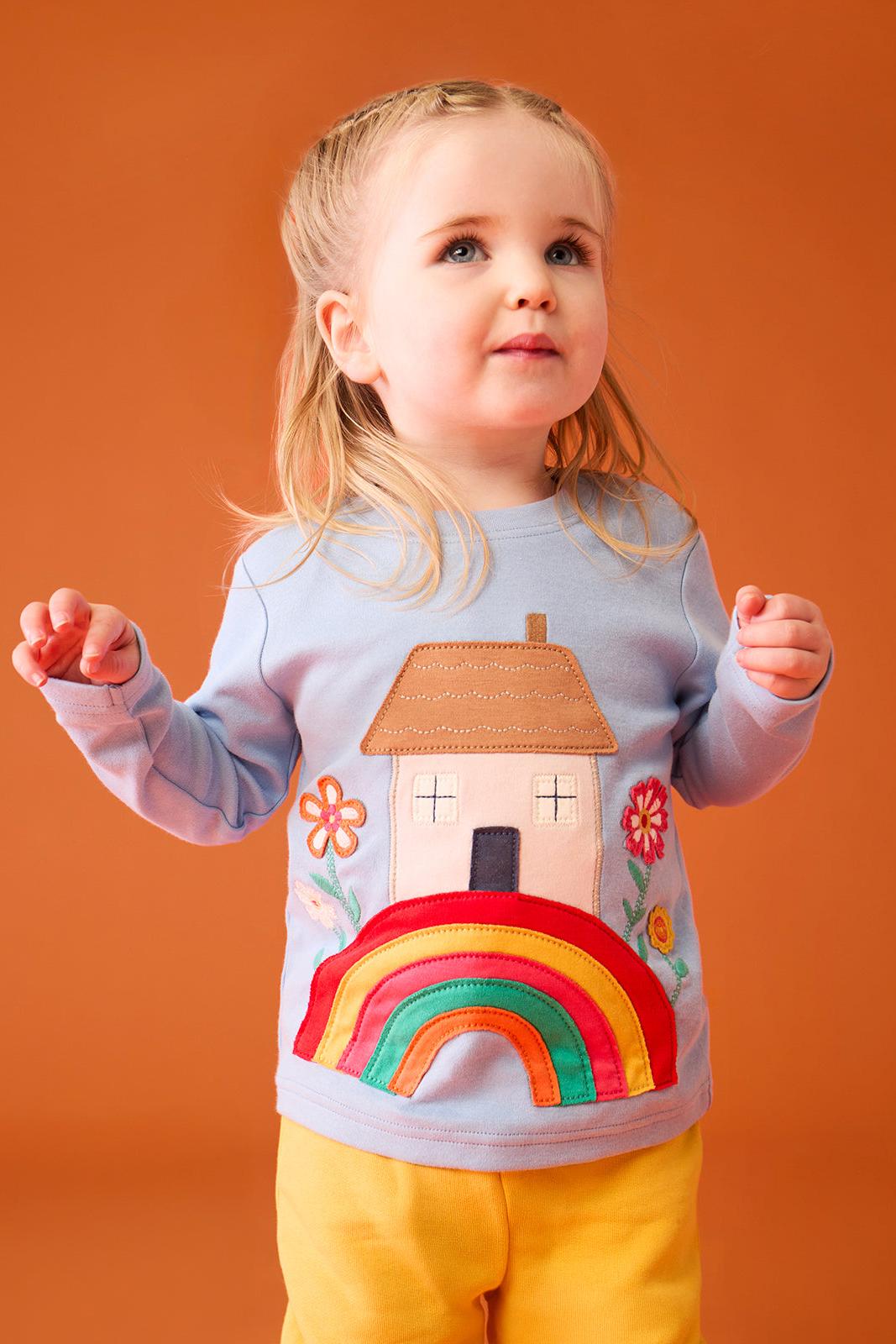 A happy toddler girl wearing a light blue Frugi organic cotton top with a rainbow and house design.