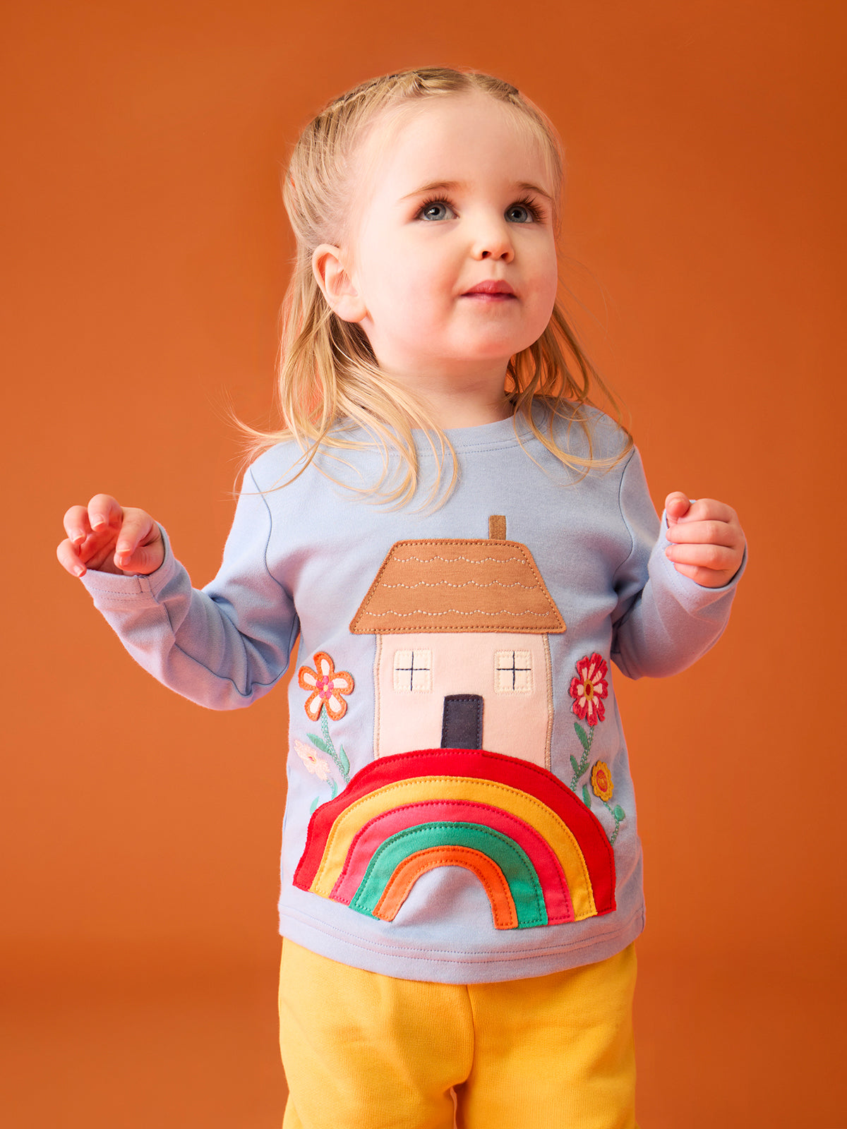 A happy toddler girl wearing a light blue Frugi organic cotton top with a rainbow and house design.