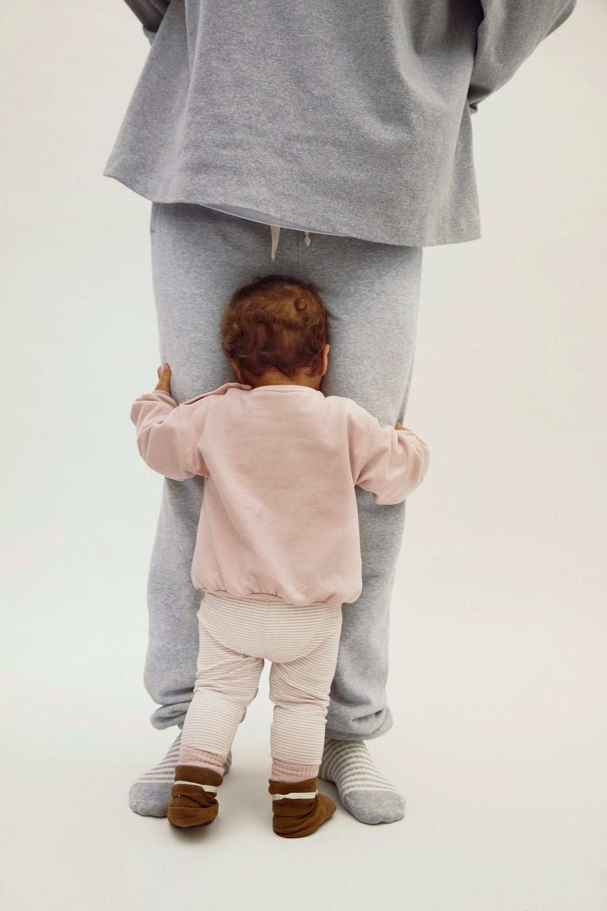 Person wearing gray pants and a pink top, with a child in a pink outfit standing behind them on a white background.