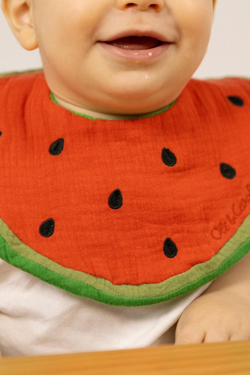 Baby wearing a watermelon-themed bib with visible brand name 'Oli&Carol'.