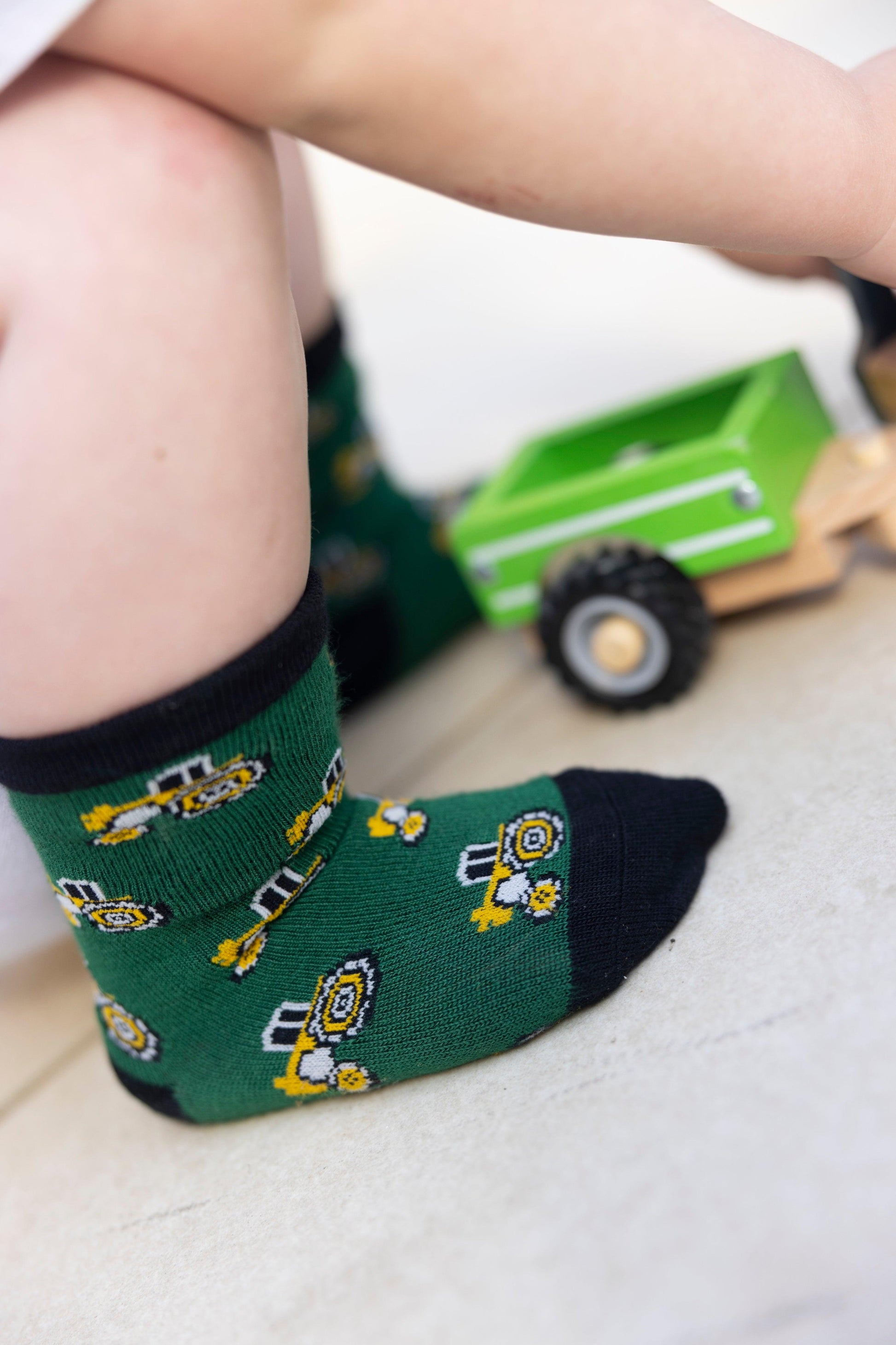 A toddler wearing green construction-themed socks playing with a wooden toy tractor on a tile floor.