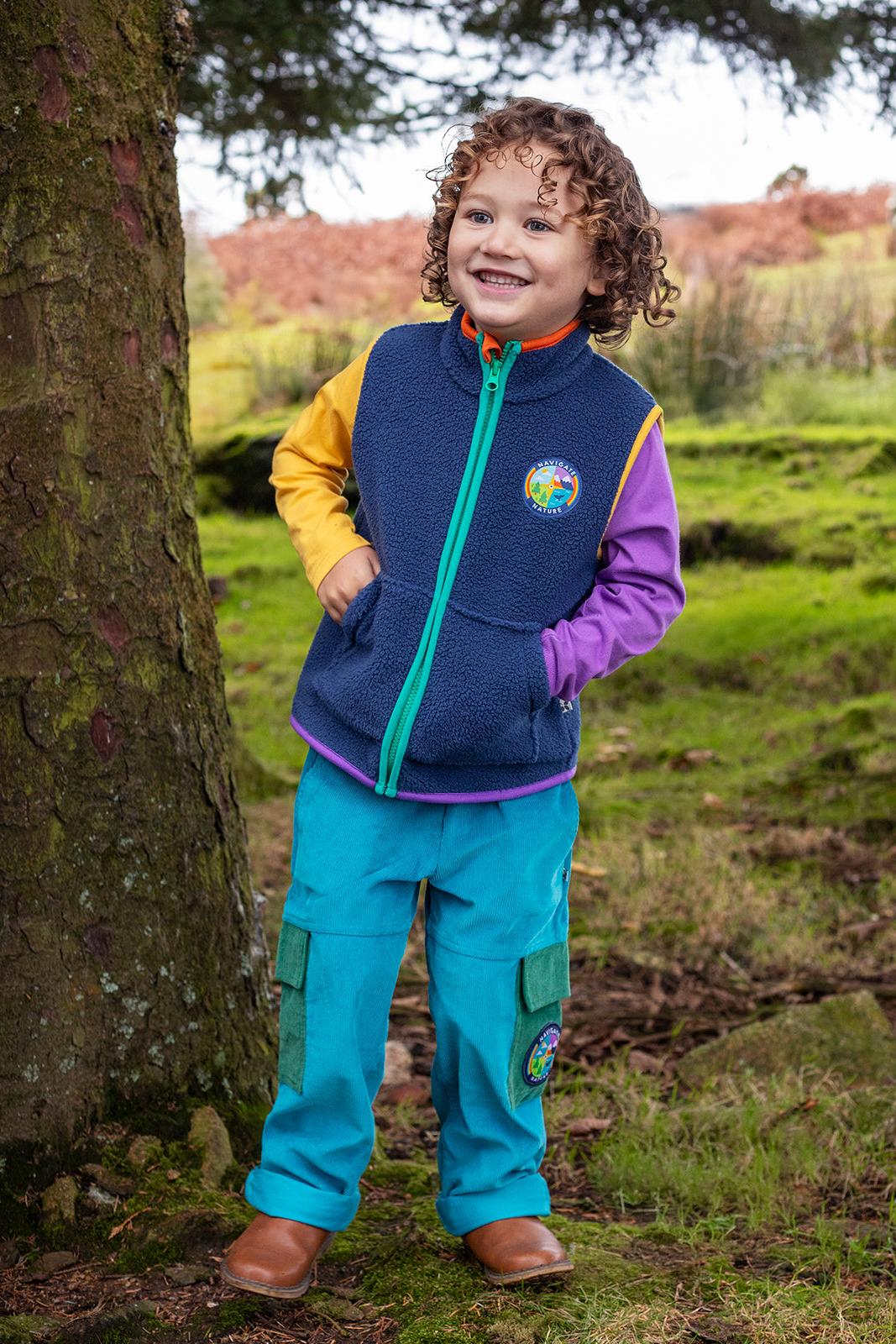 A smiling child wearing a colourful fleece vest and bright blue organic cotton cord trousers in a wooded park.