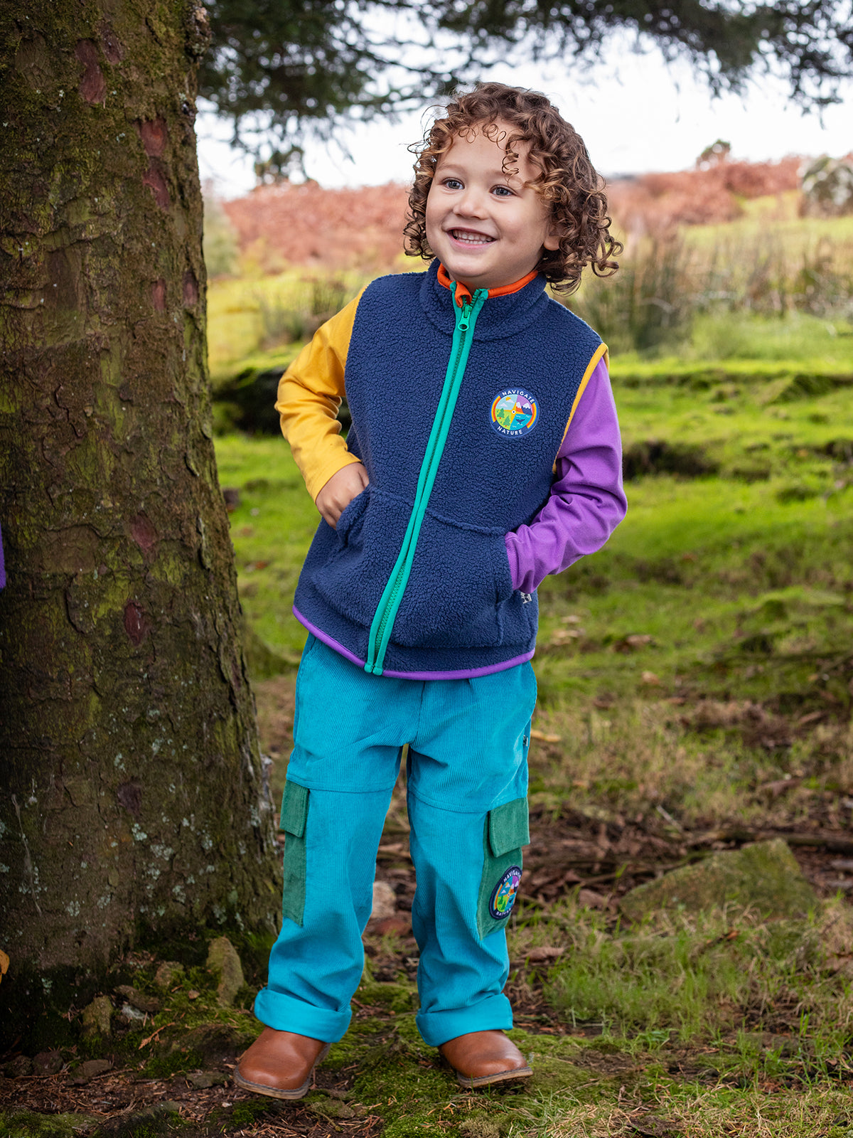 A smiling child wearing a colourful fleece vest and bright blue organic cotton cord trousers in a wooded park.