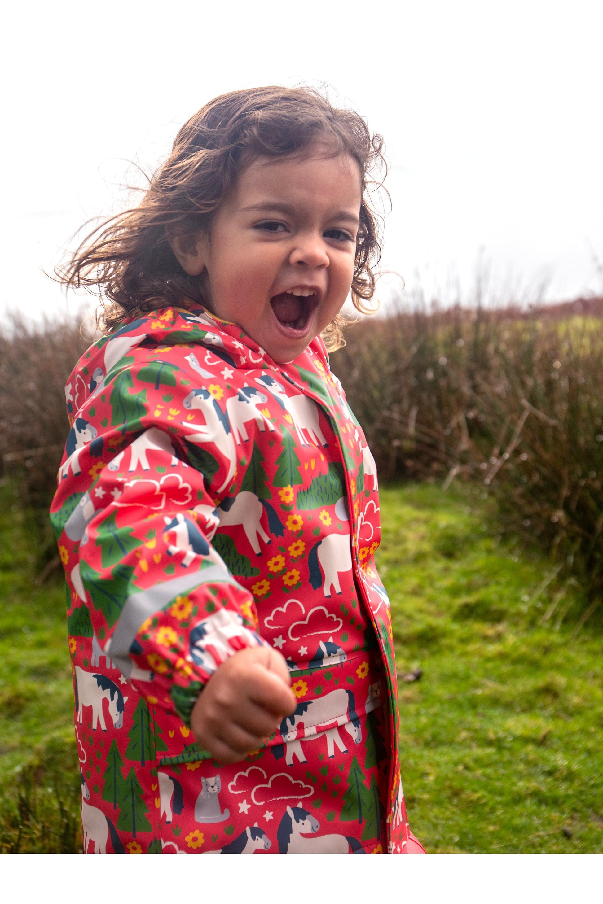 A happy toddler laughing while wearing a colorful Frugi rain jacket with horse prints in a grassy field