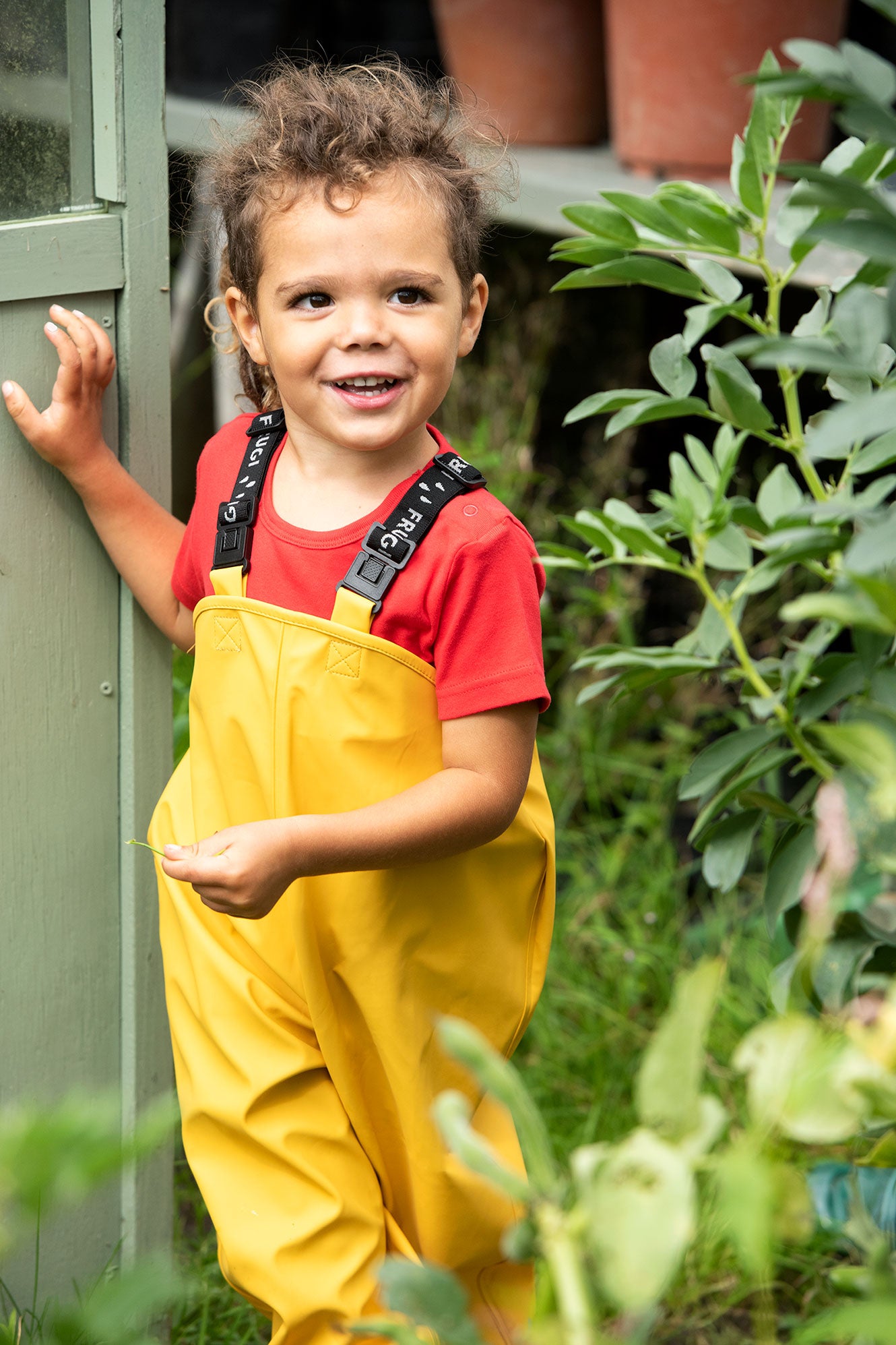 Happy toddler wearing bright yellow Frugi dungarees standing in a green garden