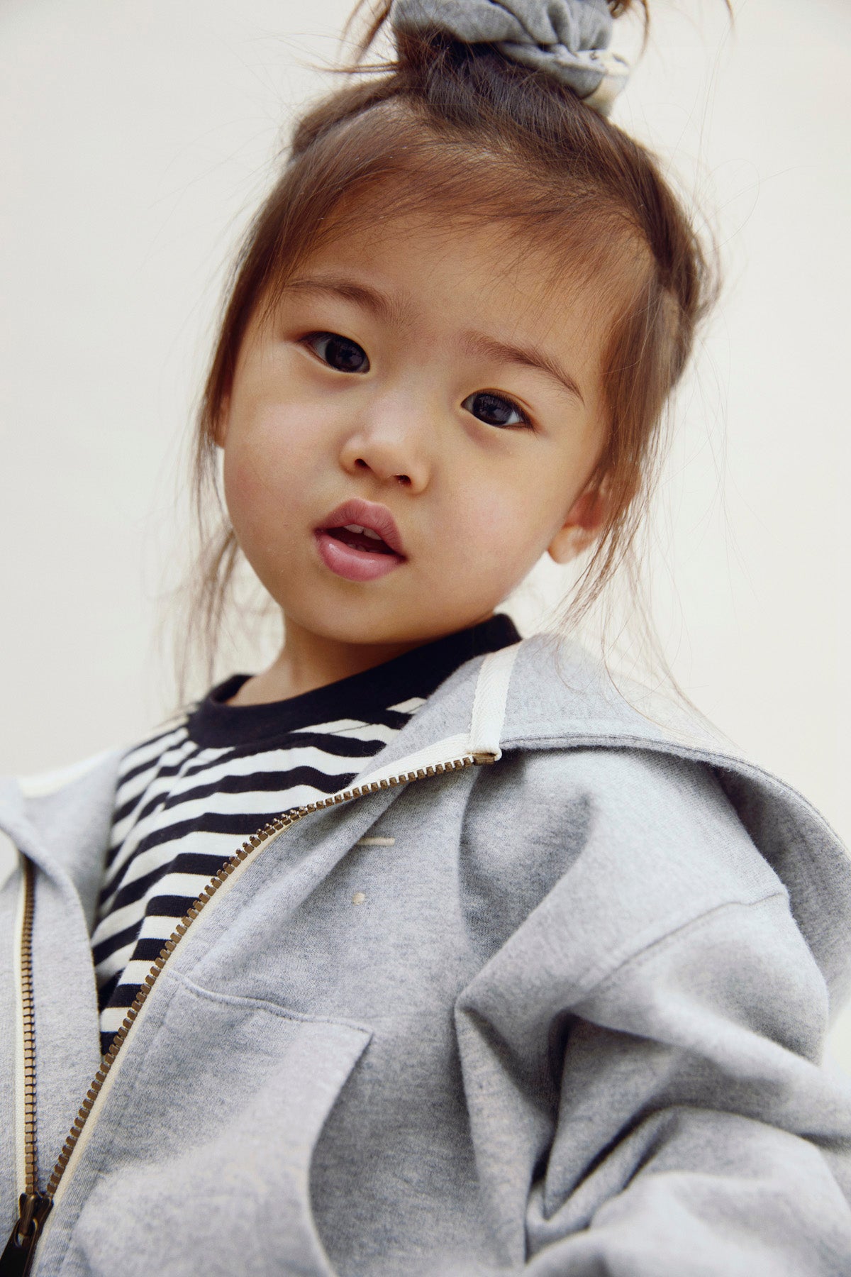 A young child wearing a light gray zip-up hoodie over a black and white striped shirt, photographed against a plain light background.