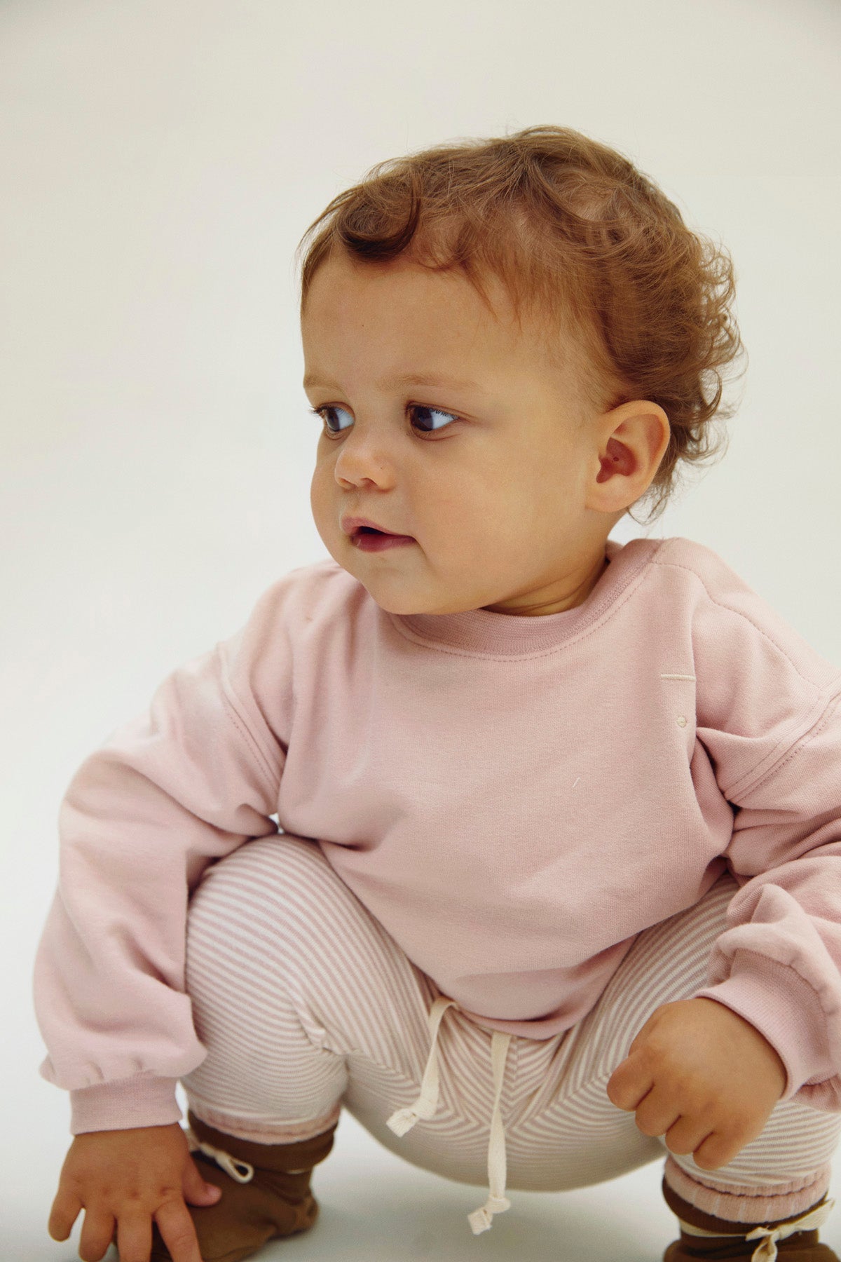 A baby wearing a light pink sweatshirt and white striped pants, crouching down against a plain light background.