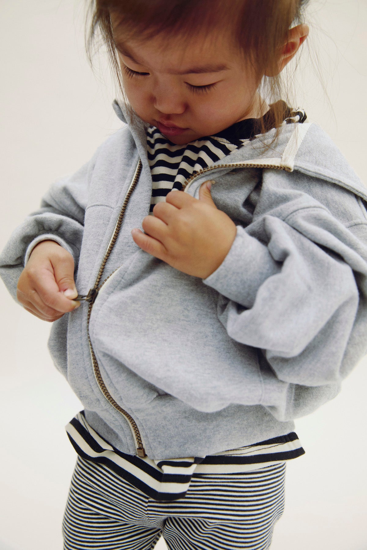 A toddler wearing a light gray zip-up hoodie over a black and white striped shirt and matching striped pants, standing against a plain light background.