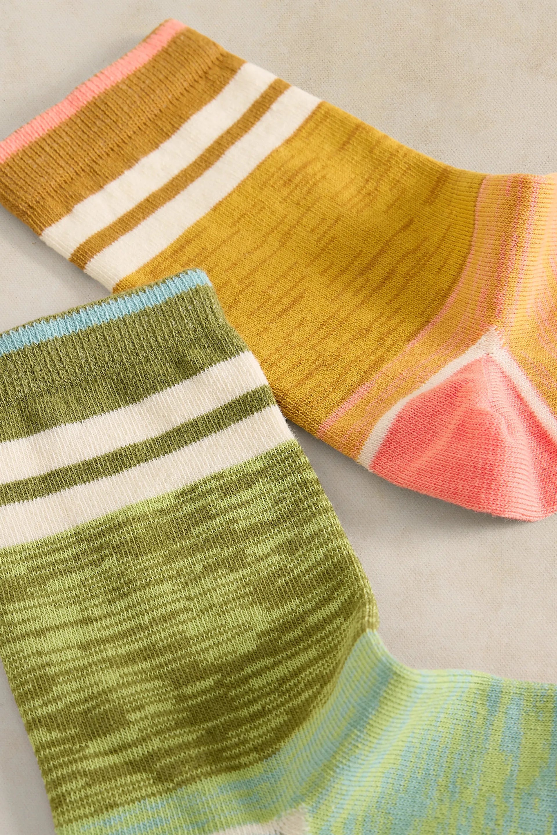 Two striped knit socks laid flat on a light beige surface. The top sock is mustard yellow with white and coral pink stripes, and the bottom sock is olive green with white and light blue stripes.