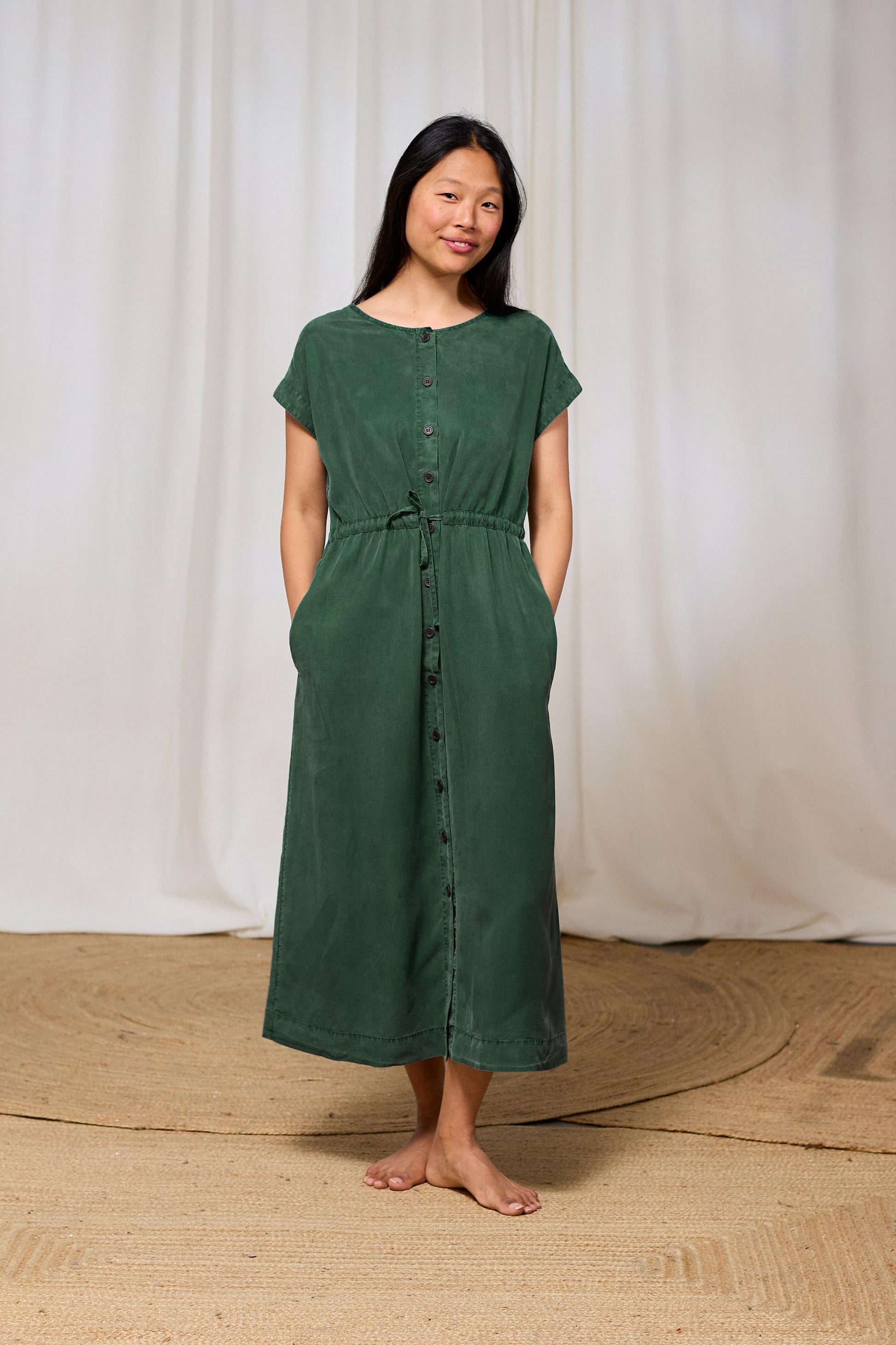 Woman wearing a green sustainable dress standing against a white curtain background
