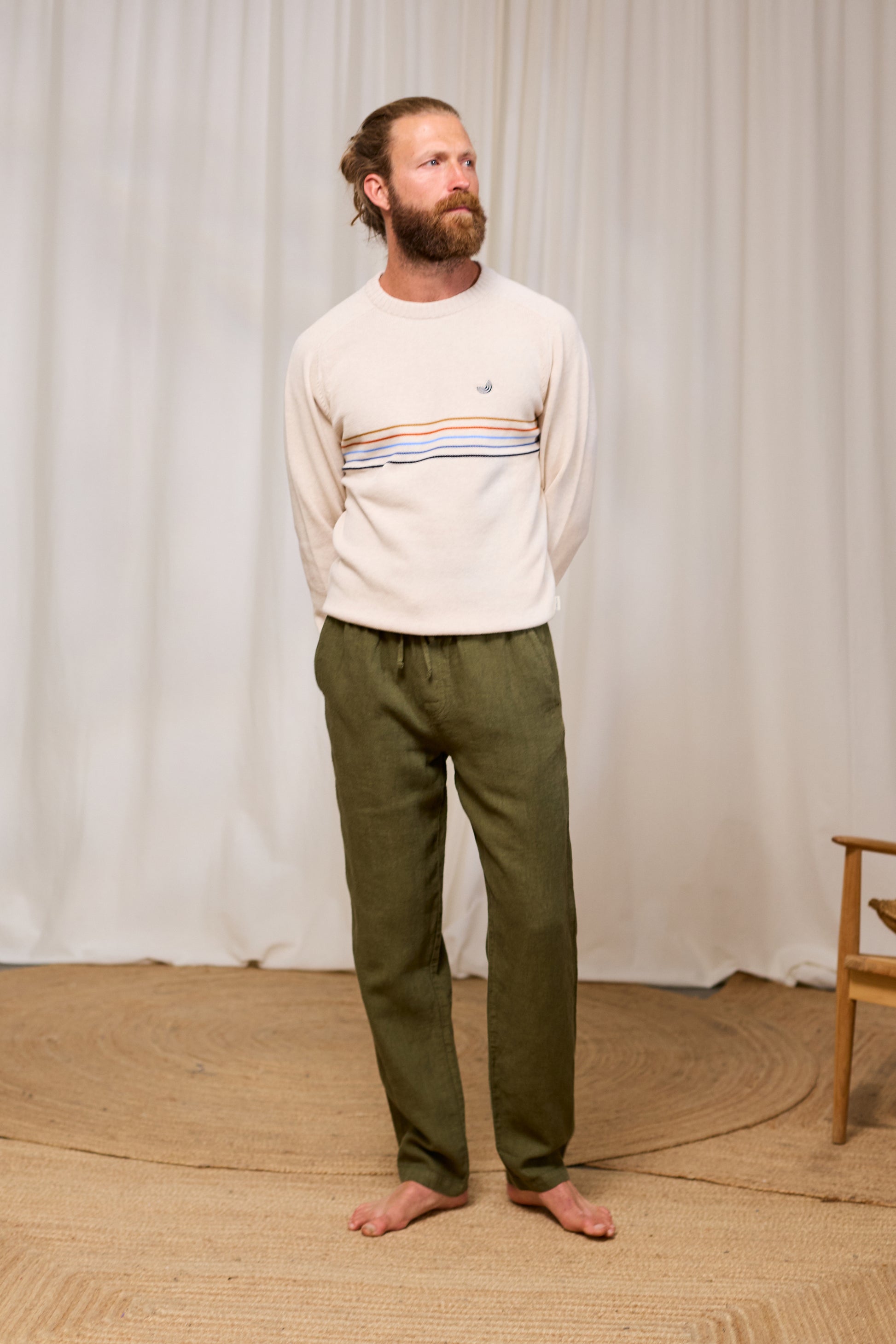 Man wearing a cream organic cotton sweater with horizontal stripes and green pants standing against a white curtain.