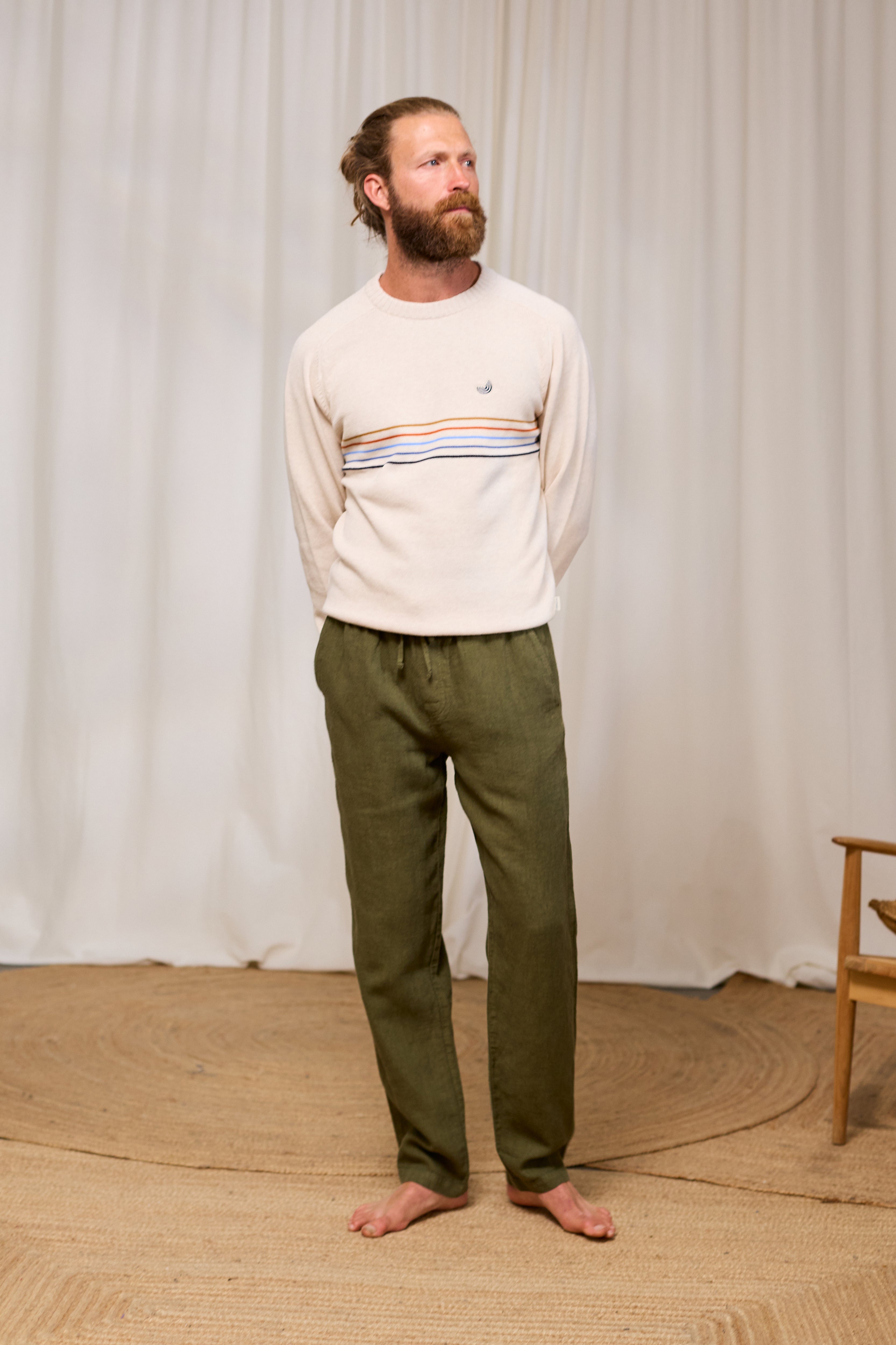 Man wearing a cream organic cotton sweater with horizontal stripes and green pants standing against a white curtain.