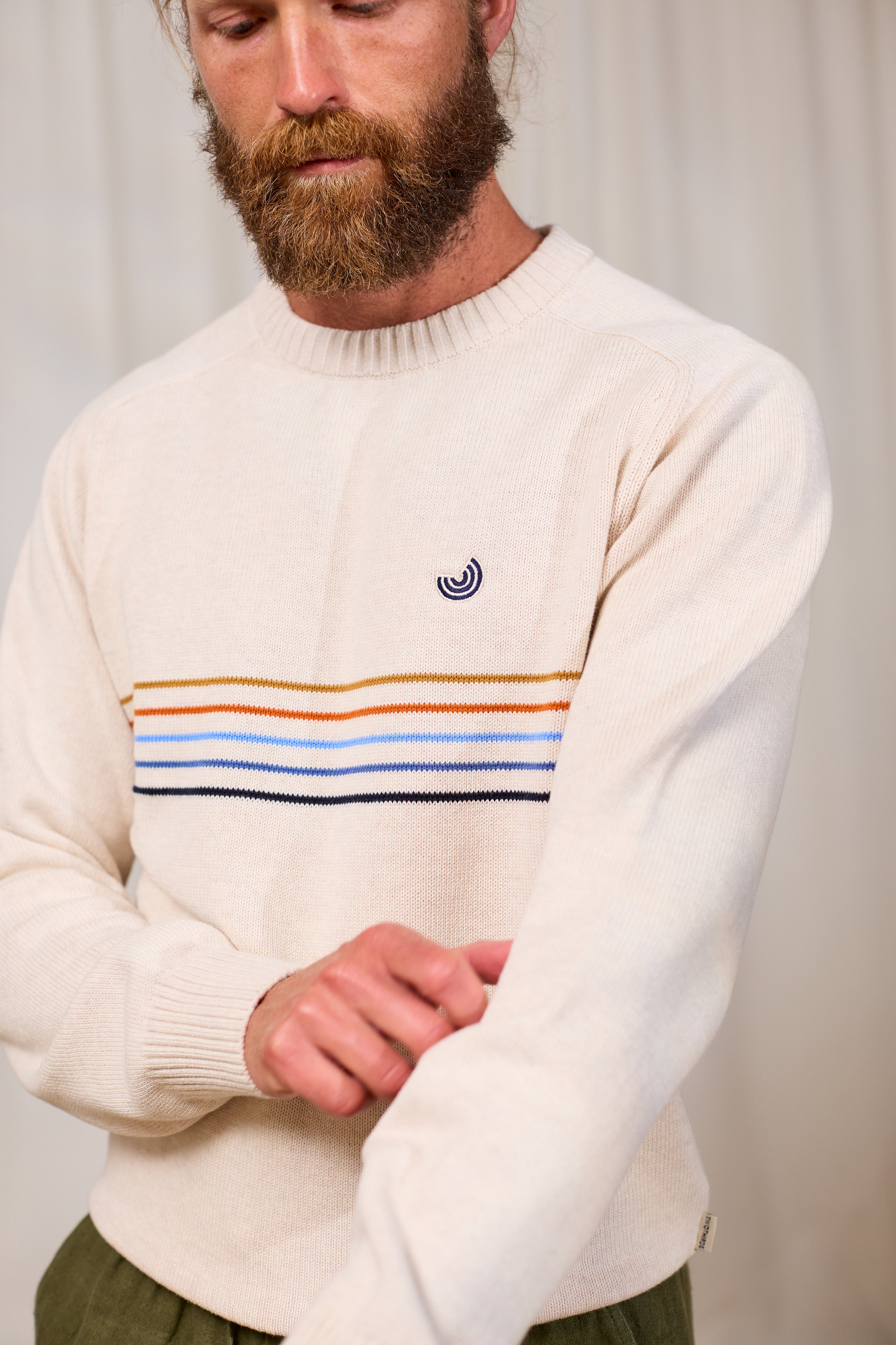 Man wearing a cream organic cotton sweater with colourful stripes and TWOTHIRDS logo, standing against a plain background