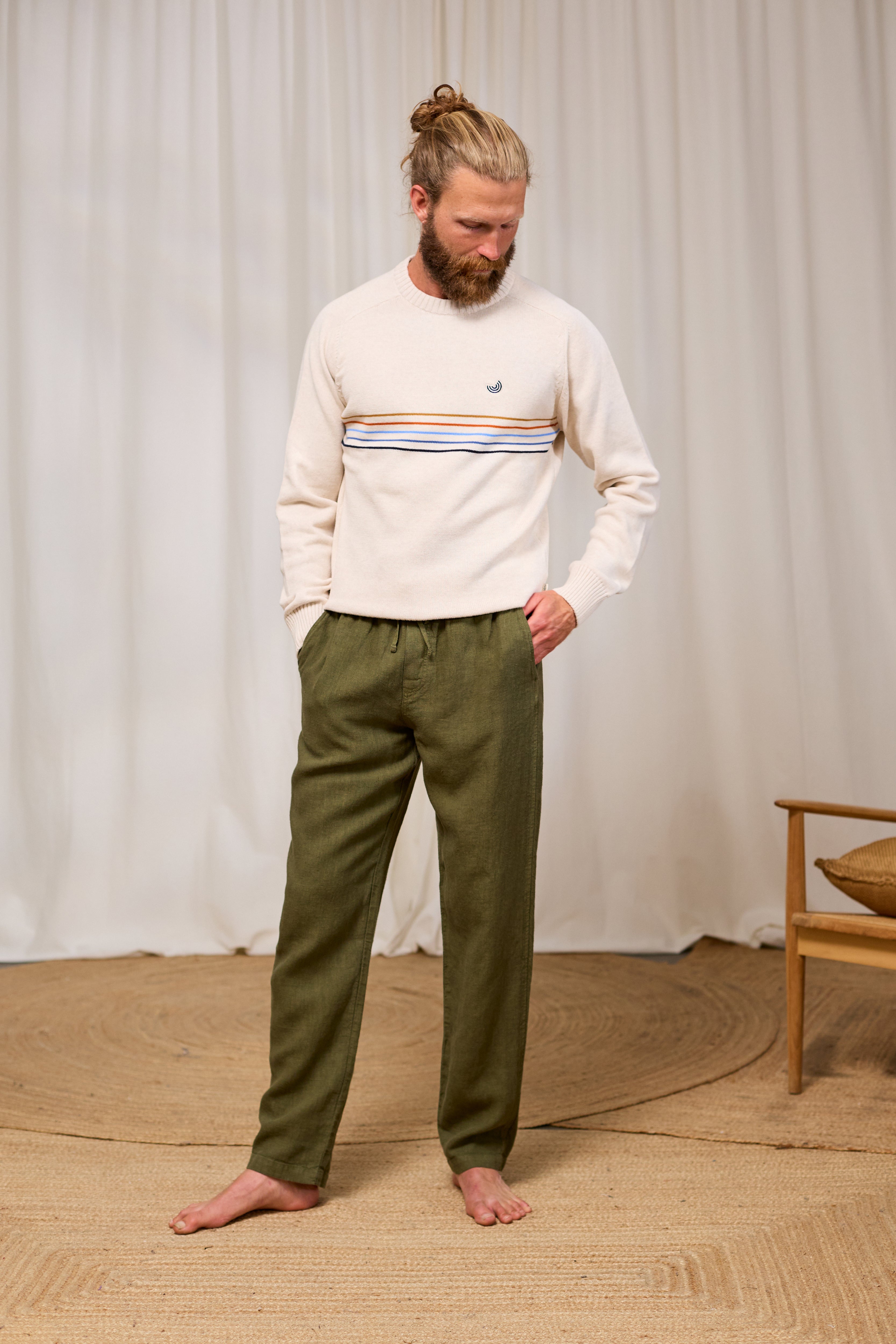 Man wearing a light-coloured TWOTHIRDS sweater with striped details and green pants standing against a white curtain.