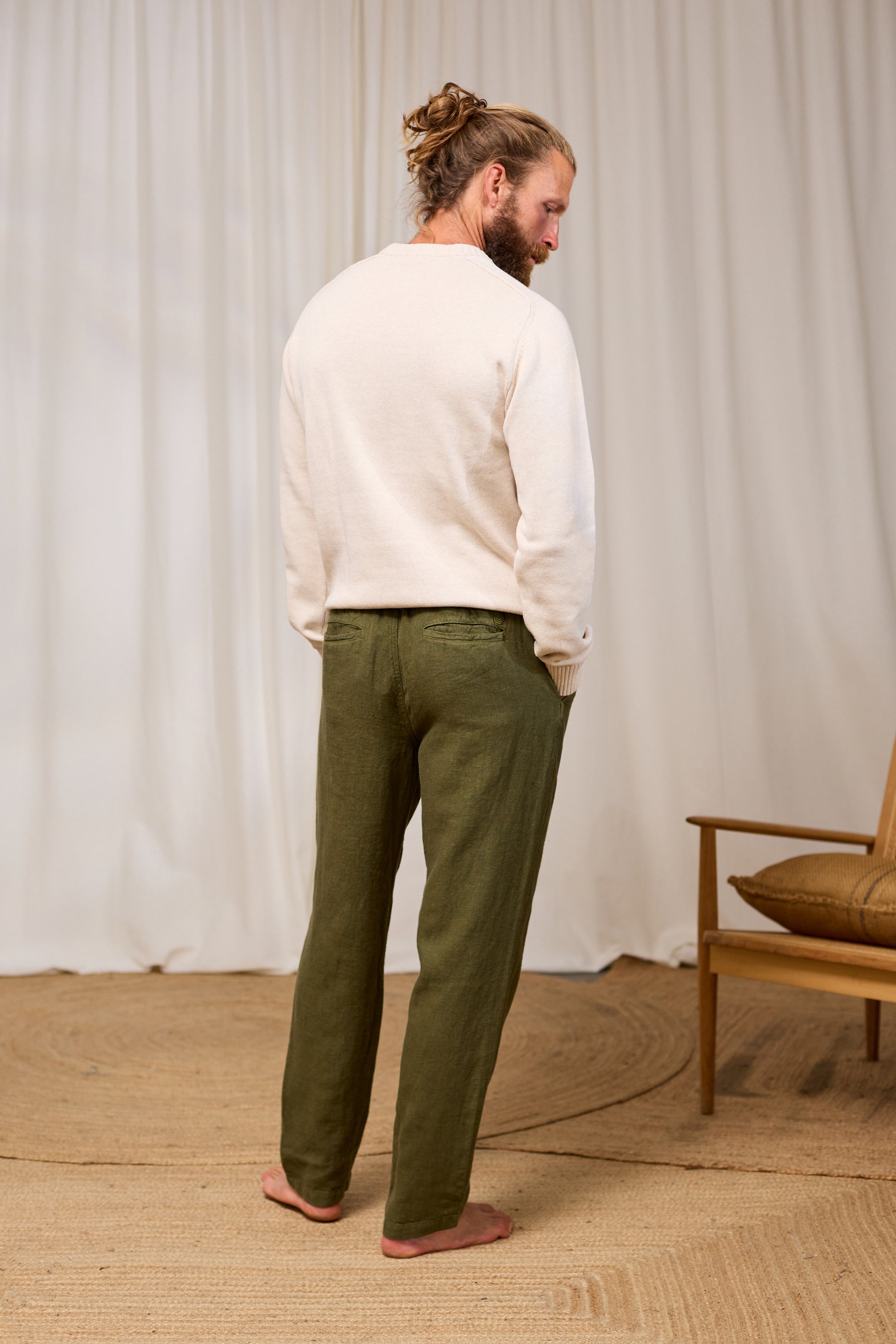 Man wearing an ecru sweater and green pants standing in a room with a wooden chair and beige rug.
