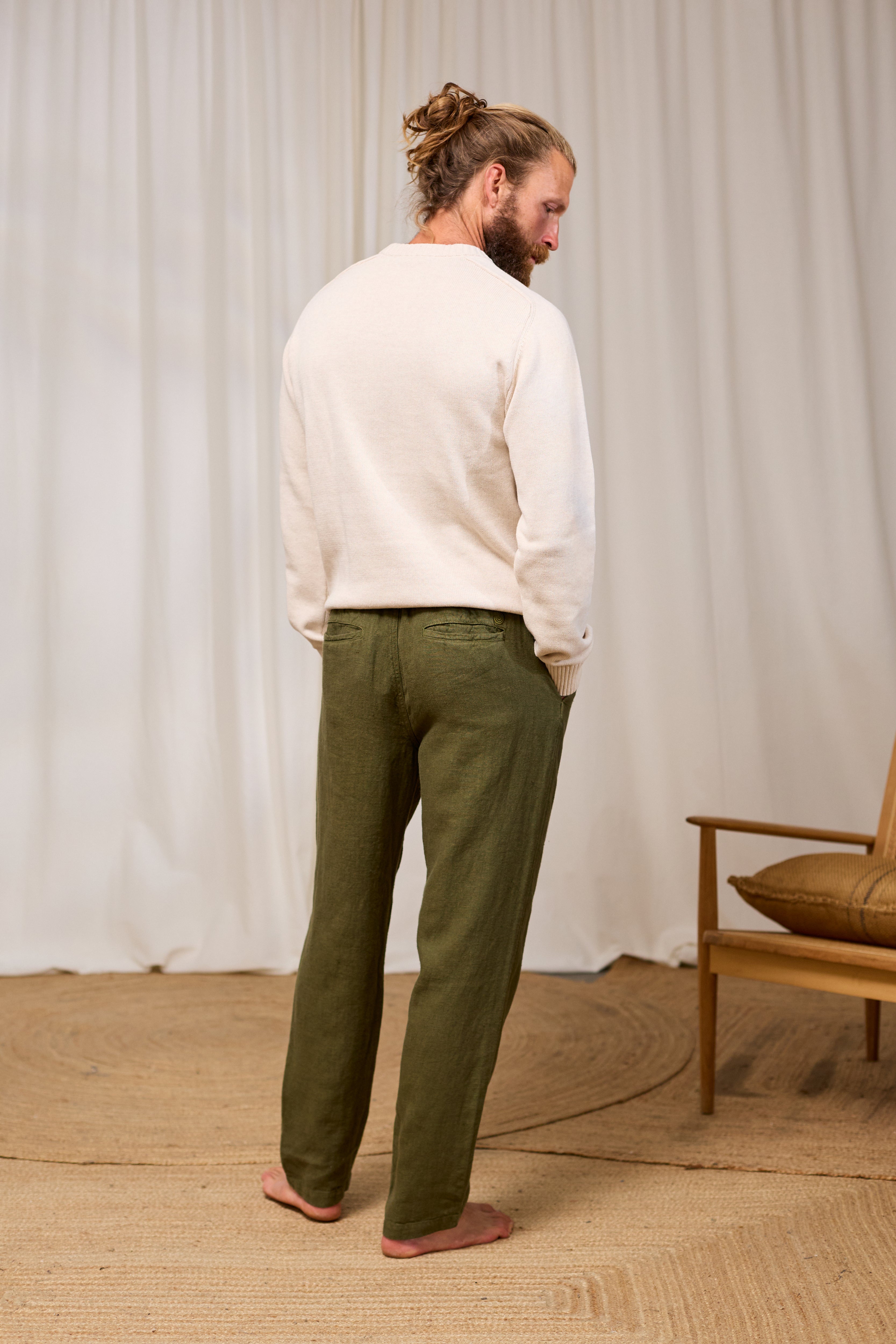Man wearing an ecru sweater and green pants standing in a room with a wooden chair and beige rug.