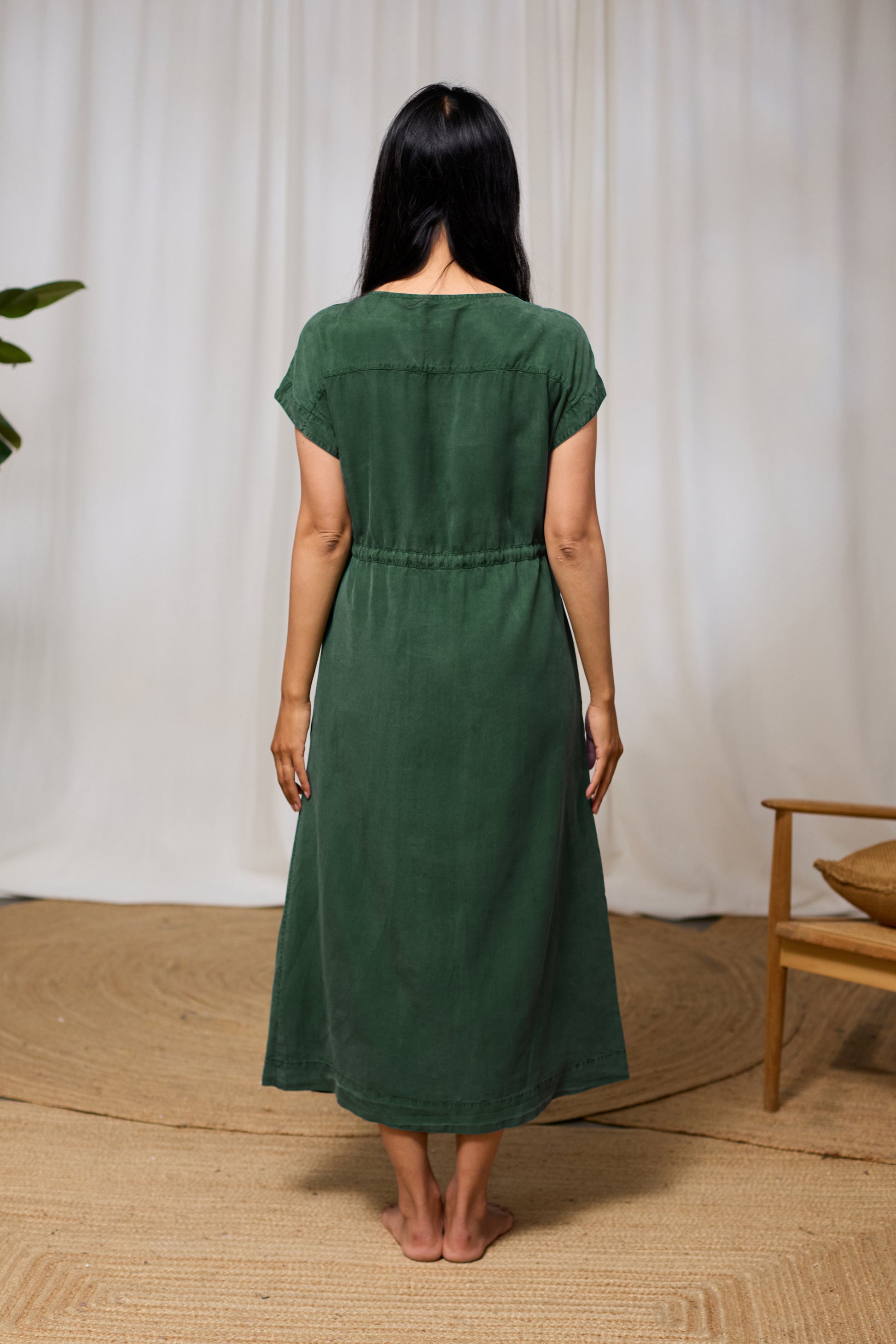 Woman wearing a green tencel dress standing in a room with a white curtain and wooden chair.