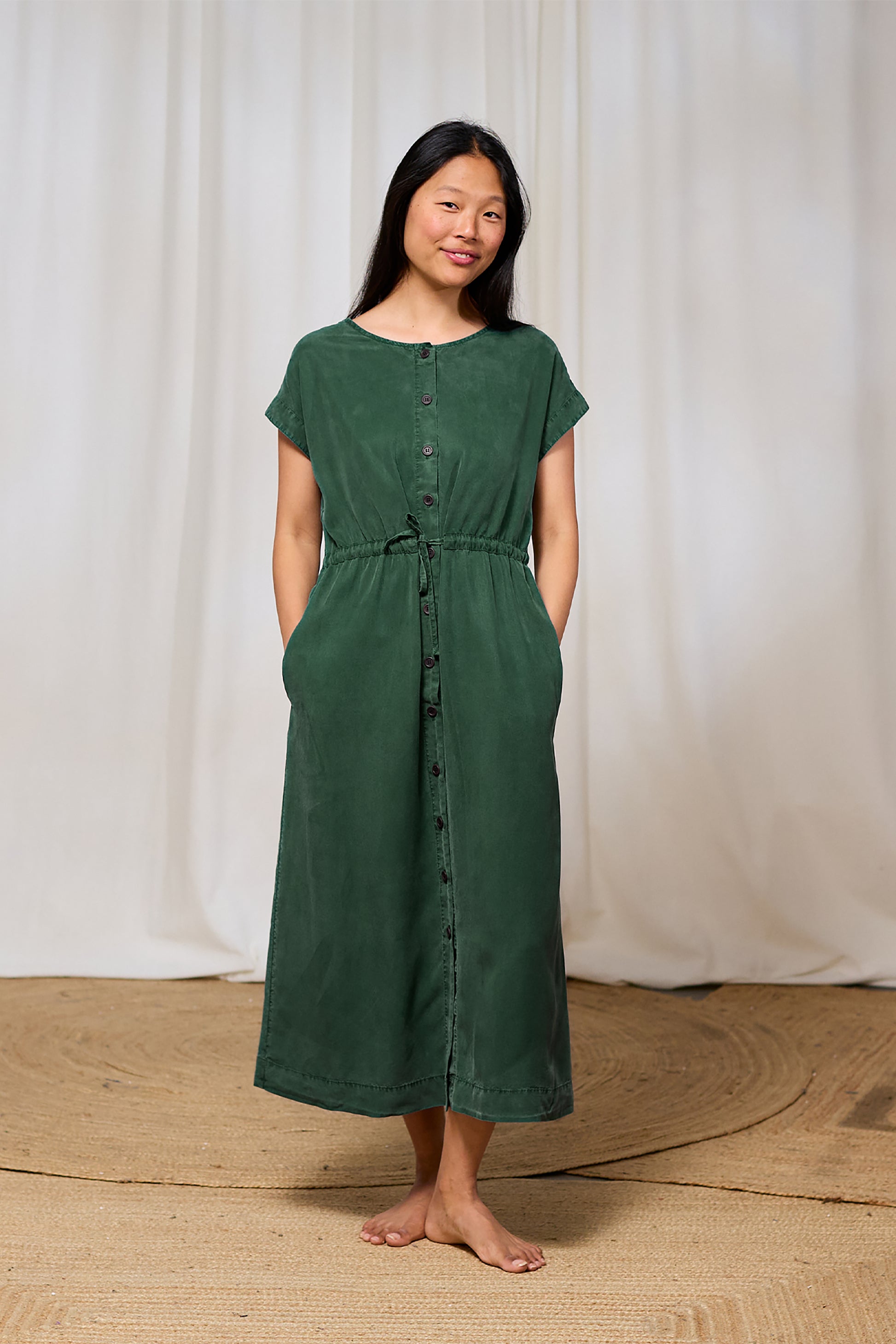 Woman wearing a green tencel dress standing against a white curtain background
