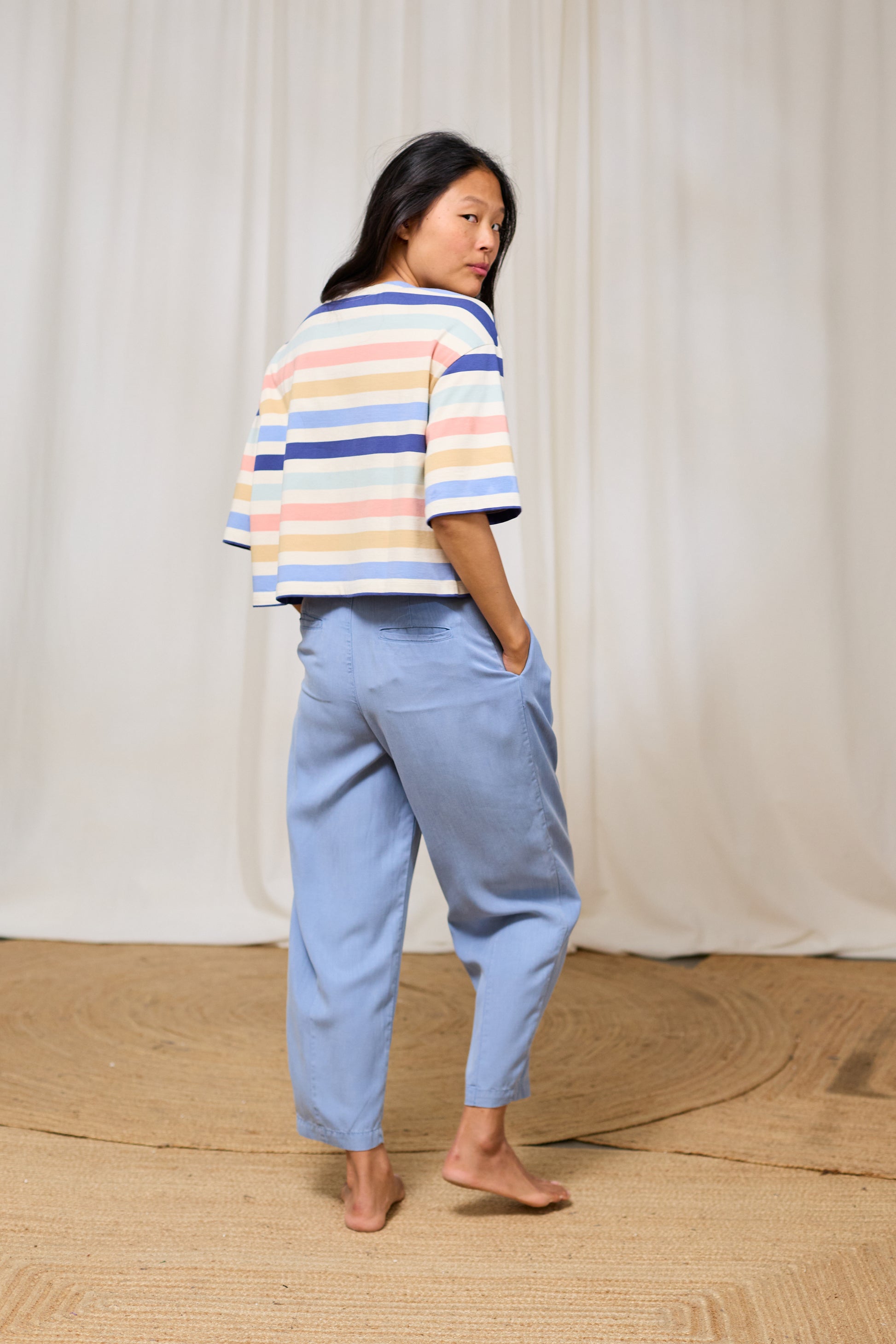 Woman wearing a striped organic cotton t-shirt and light blue pants against a white curtain background