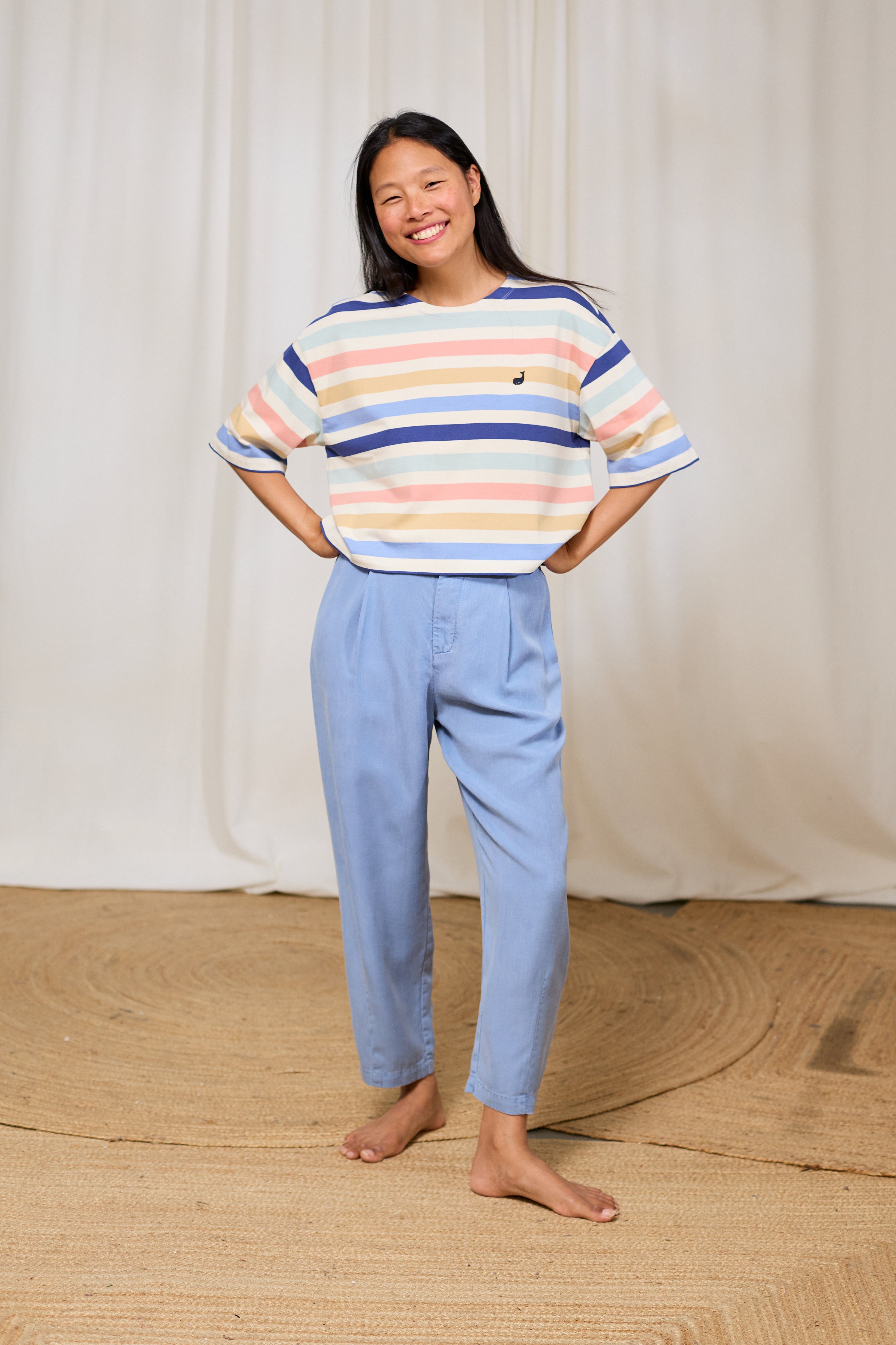 Woman wearing a colorful striped organic cotton top and light blue pants standing against a white curtain.