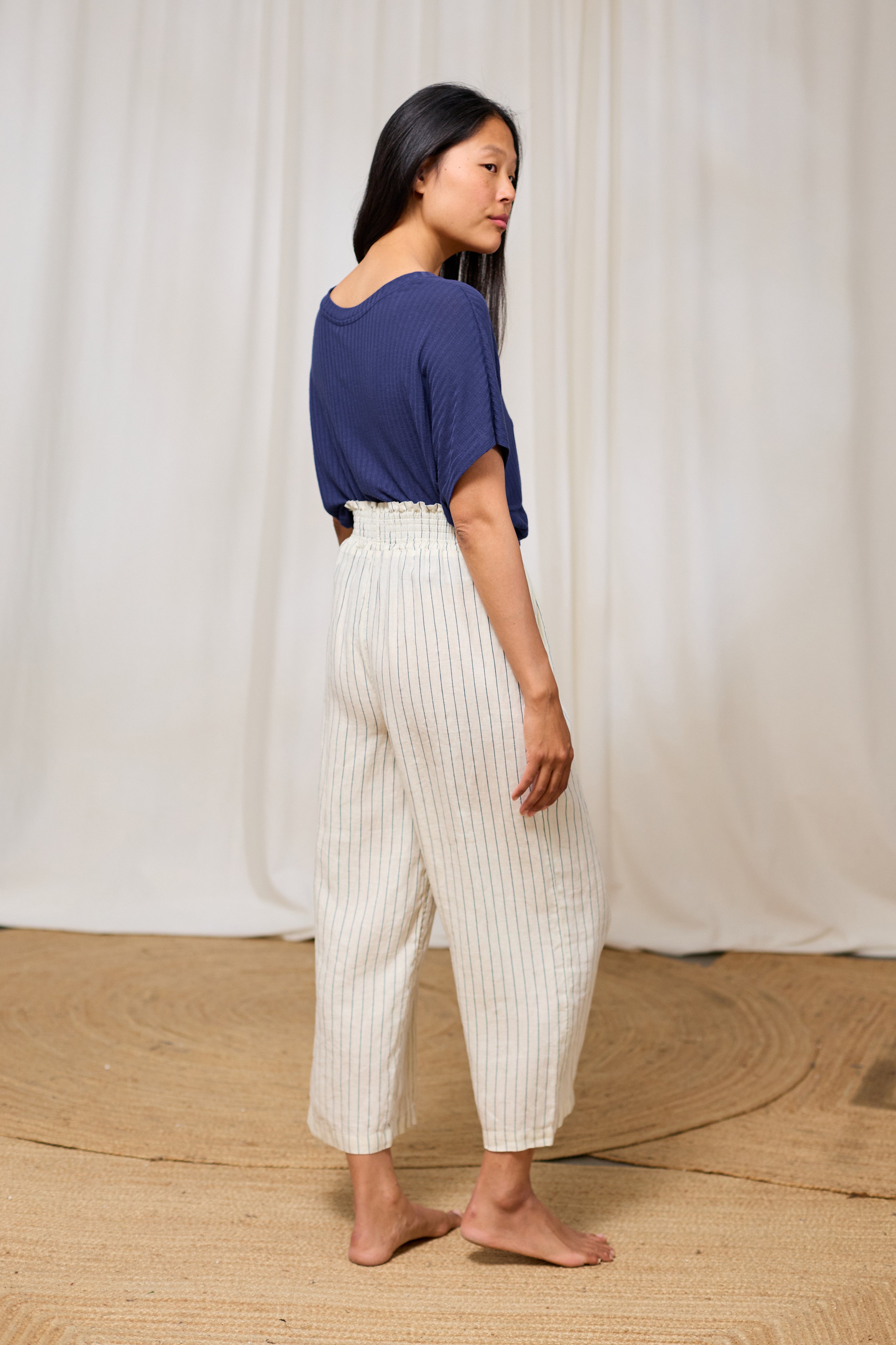 Woman wearing a blue TWOTHIRDS top and white pants standing against a white curtain.