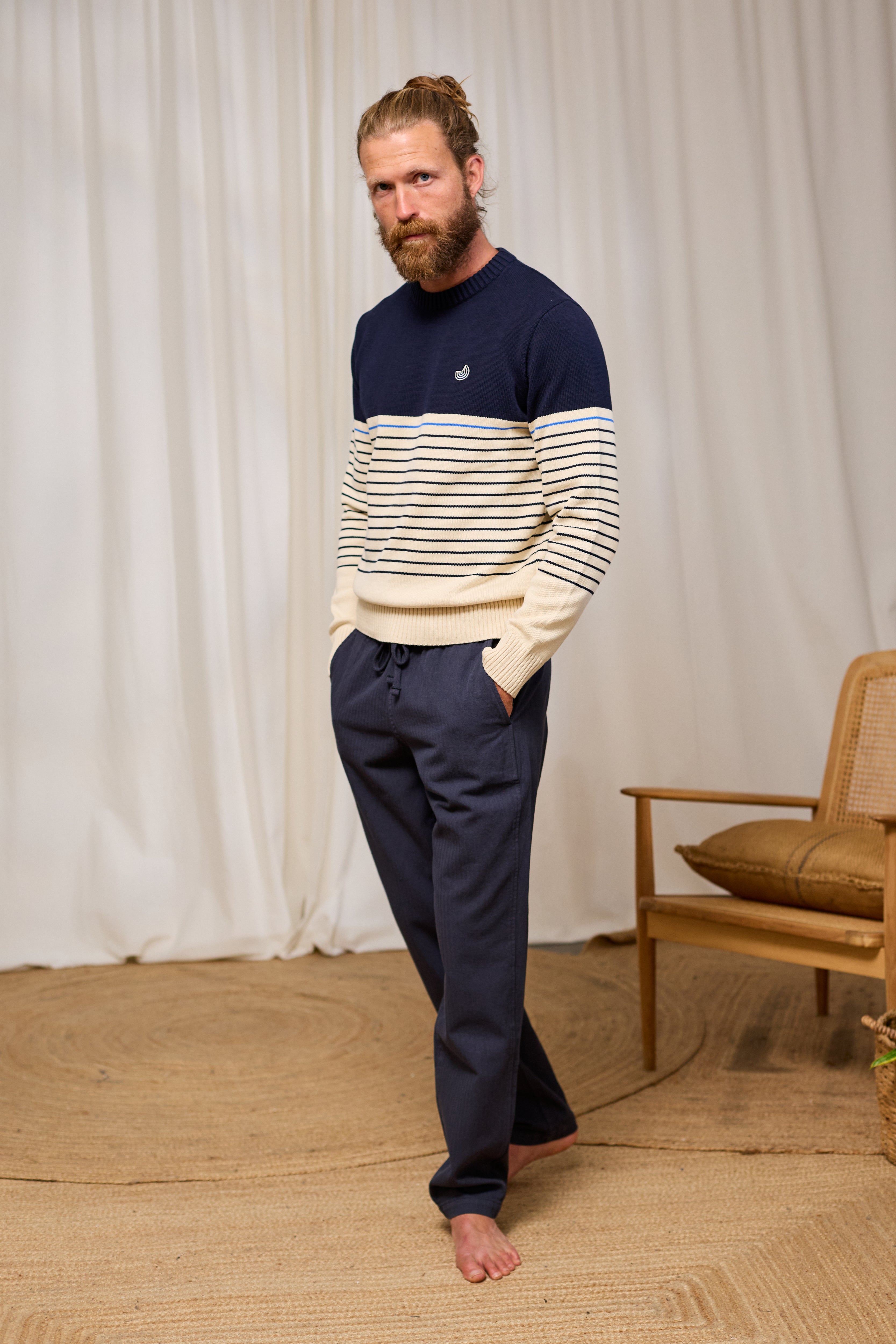 Man wearing a navy and white striped organic cotton sweater with dark pants standing in a room with a wooden chair and white curtains.