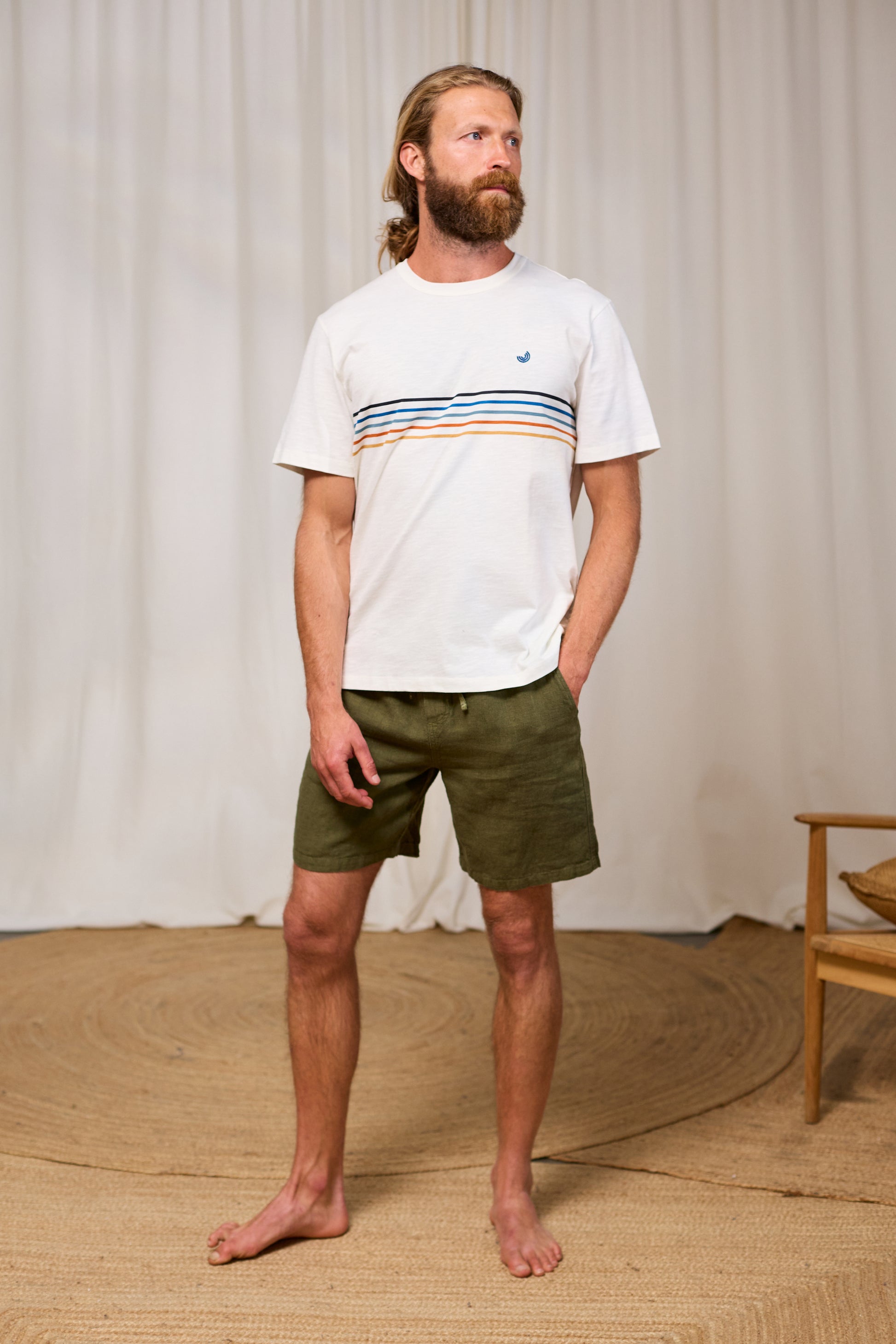 Man wearing a white roundneck t-shirt with colorful stripes and green shorts standing against a plain background.