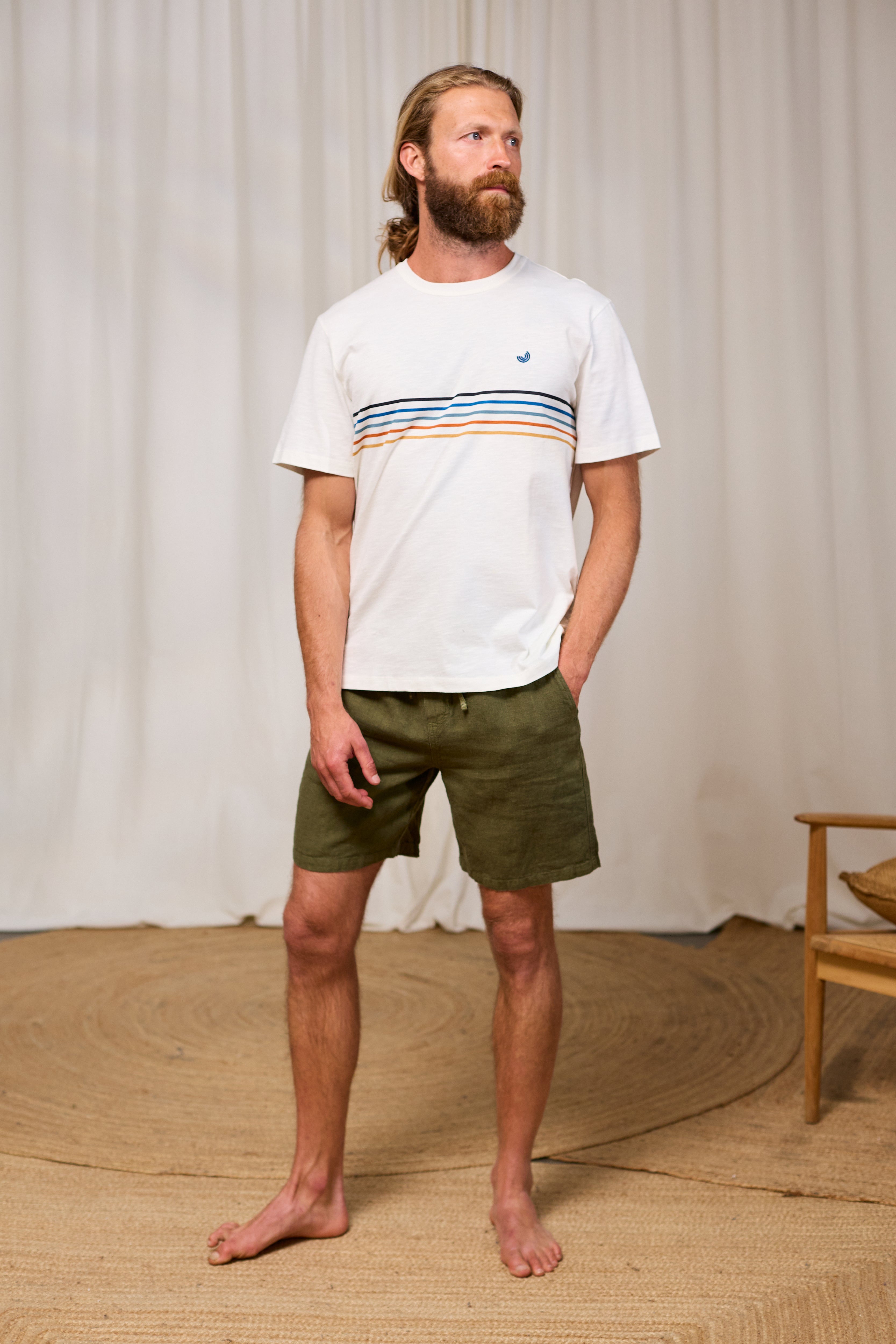Man wearing a white roundneck t-shirt with colorful stripes and green shorts standing against a plain background.