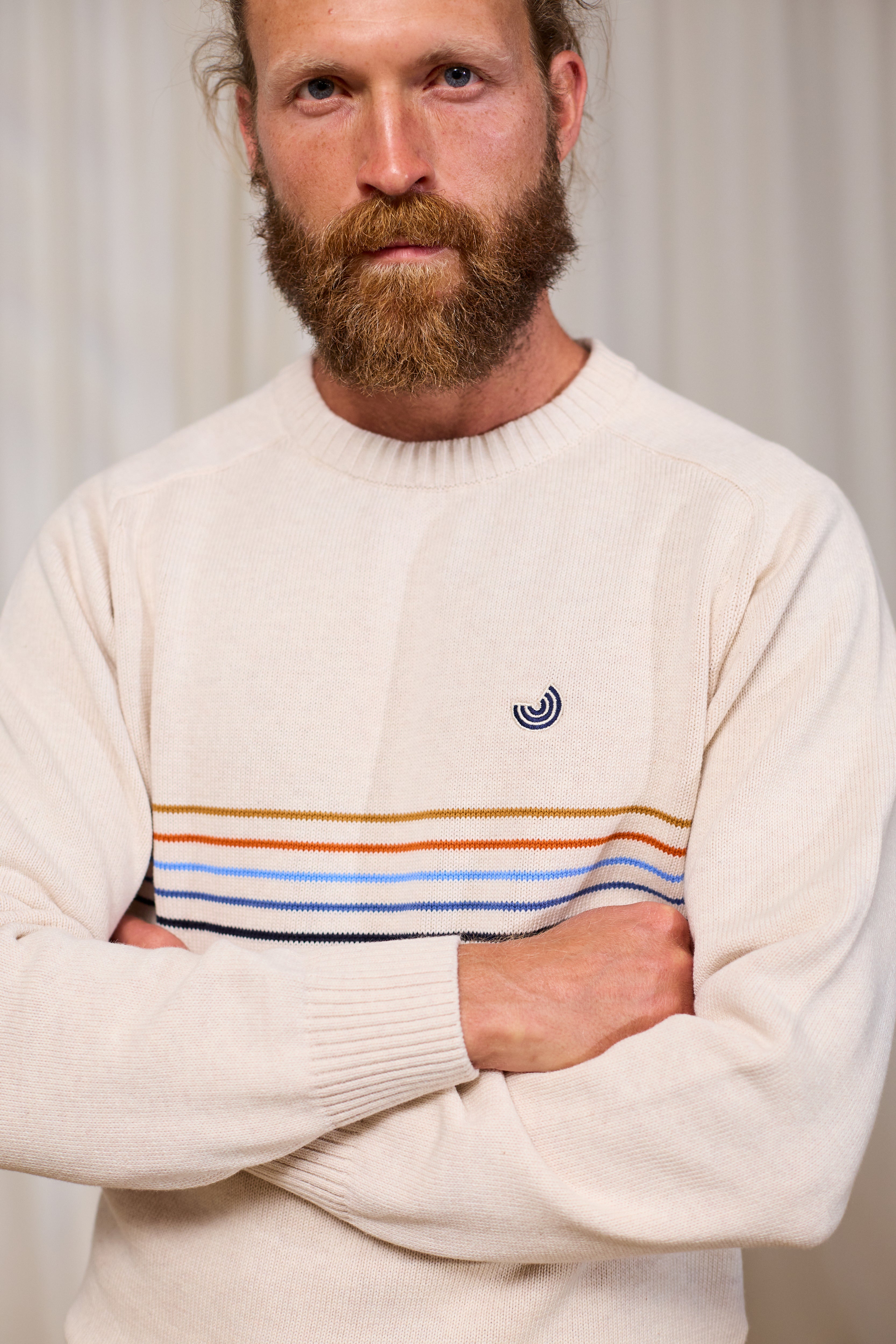 Man wearing a cream organic cotton sweater with colourful stripes and a logo, standing against a plain background.