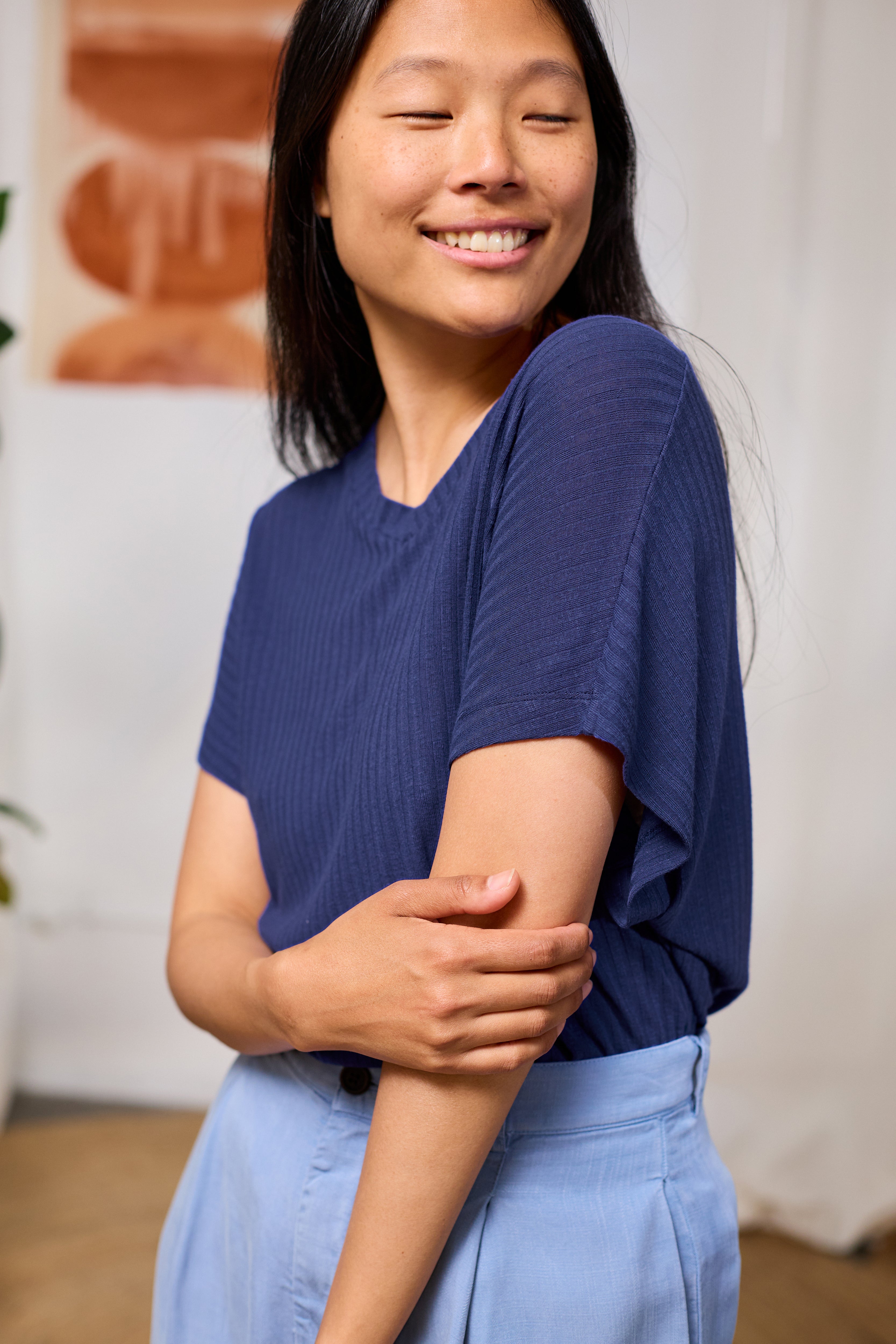 Woman wearing a navy blue ribbed top and light blue pants smiling indoors.