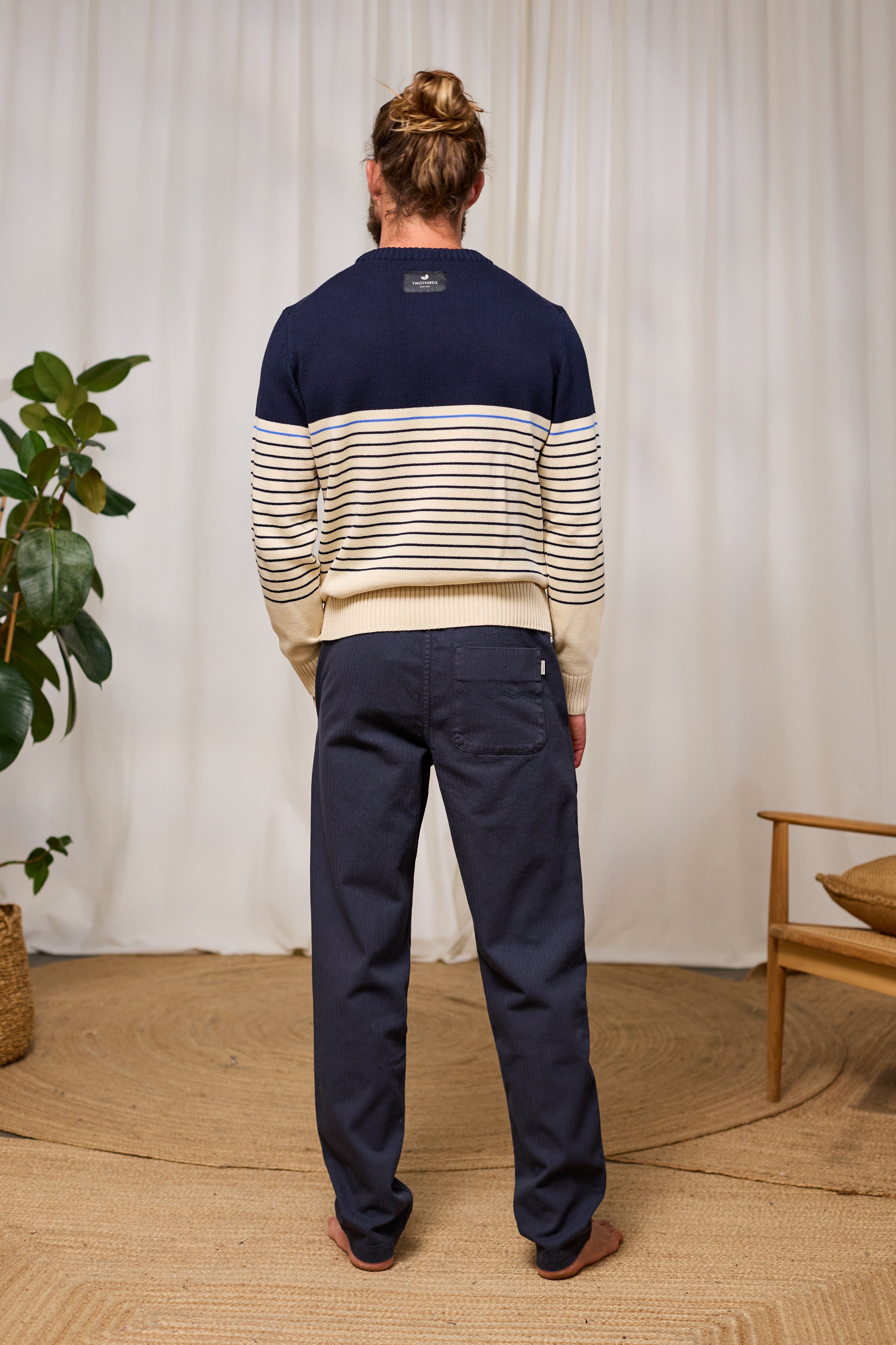 Person wearing a navy and beige striped sweater with navy pants in a room with a plant and chair.