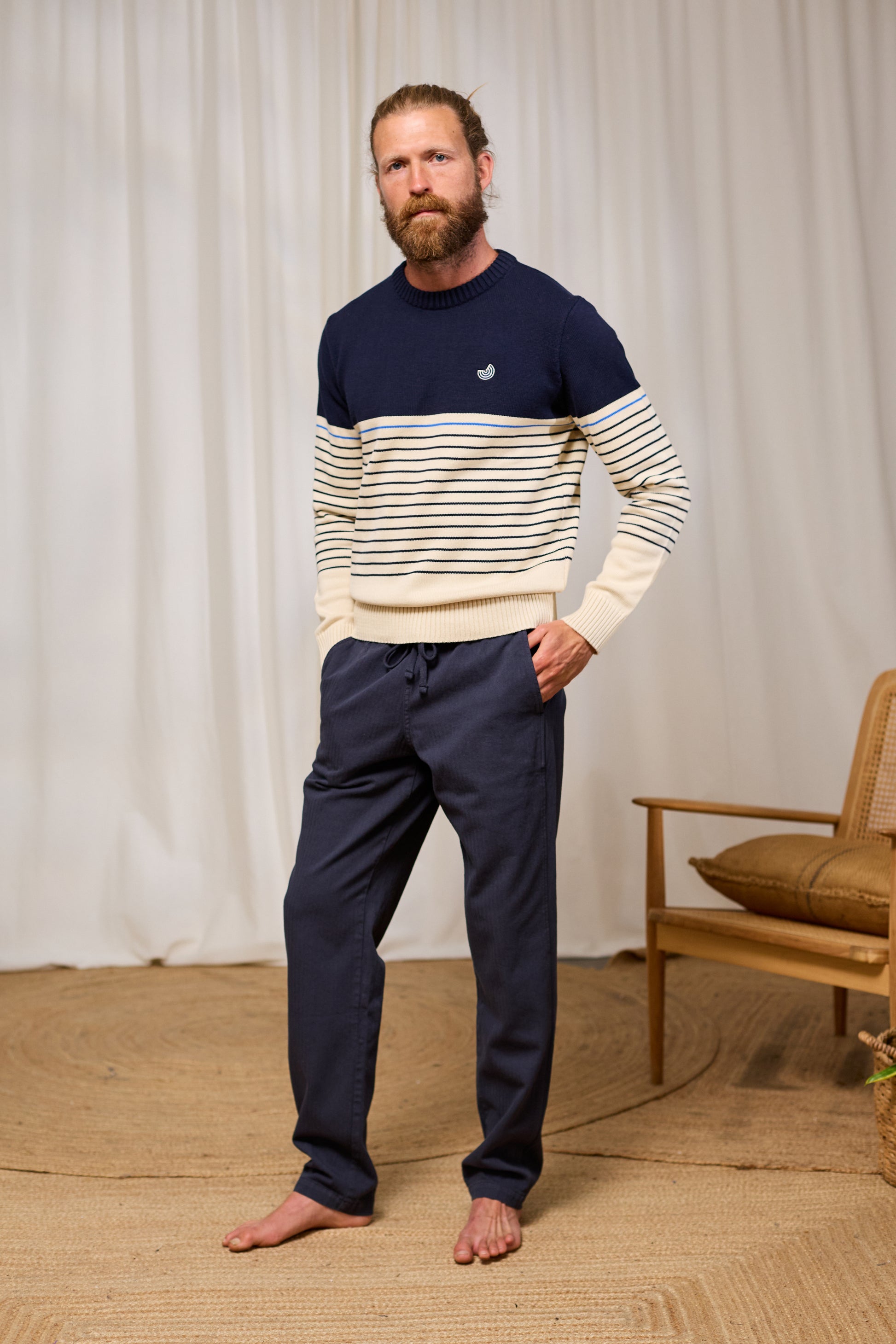 Man wearing a navy and white striped organic cotton sweater with dark pants standing in a room with a wooden chair and beige wall.