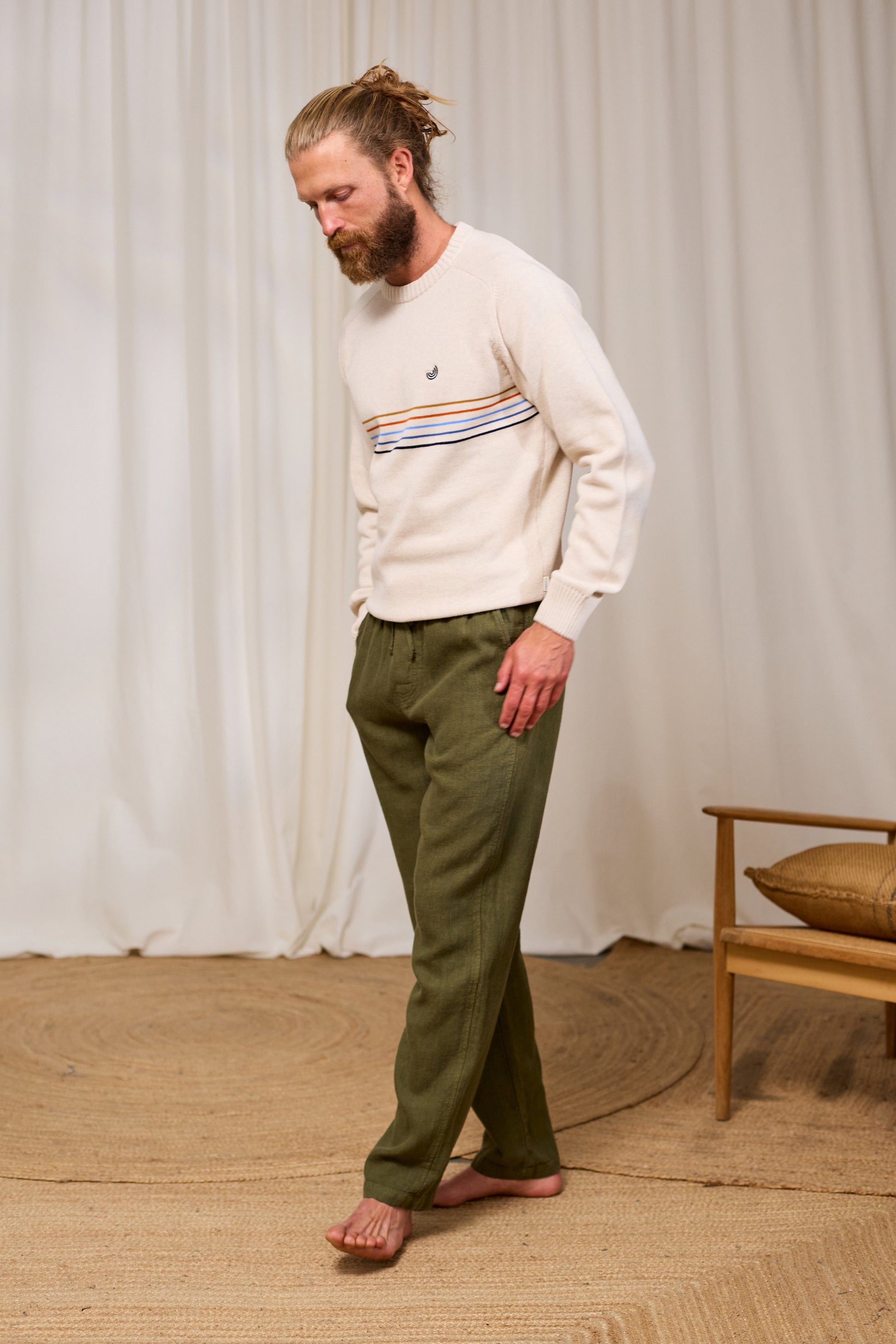 Man wearing a cream organic cotton sweater with a pattern and green pants standing in a room with a white curtain and wooden chair.