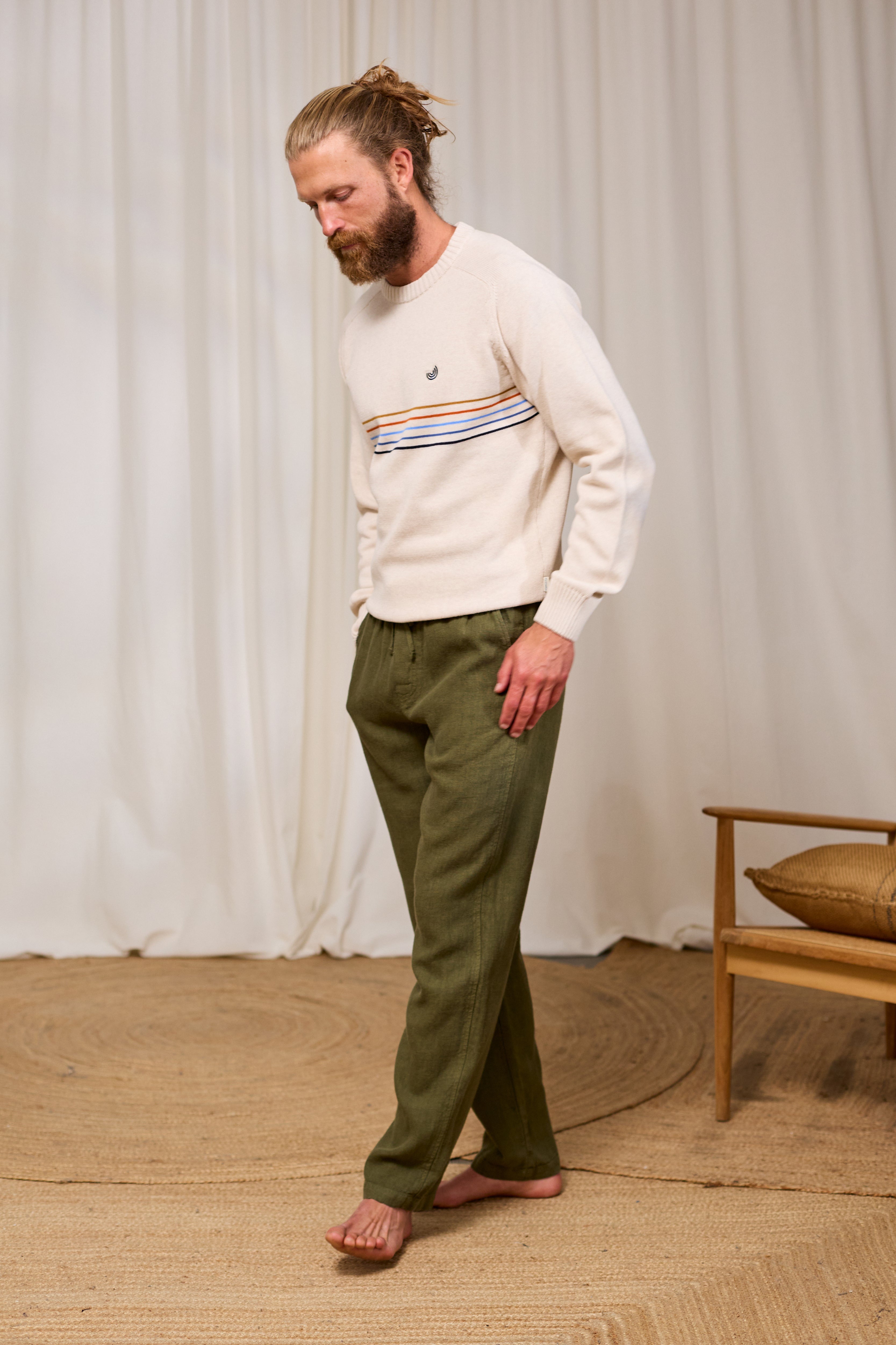 Man wearing a cream organic cotton sweater with a pattern and green pants standing in a room with a white curtain and wooden chair.