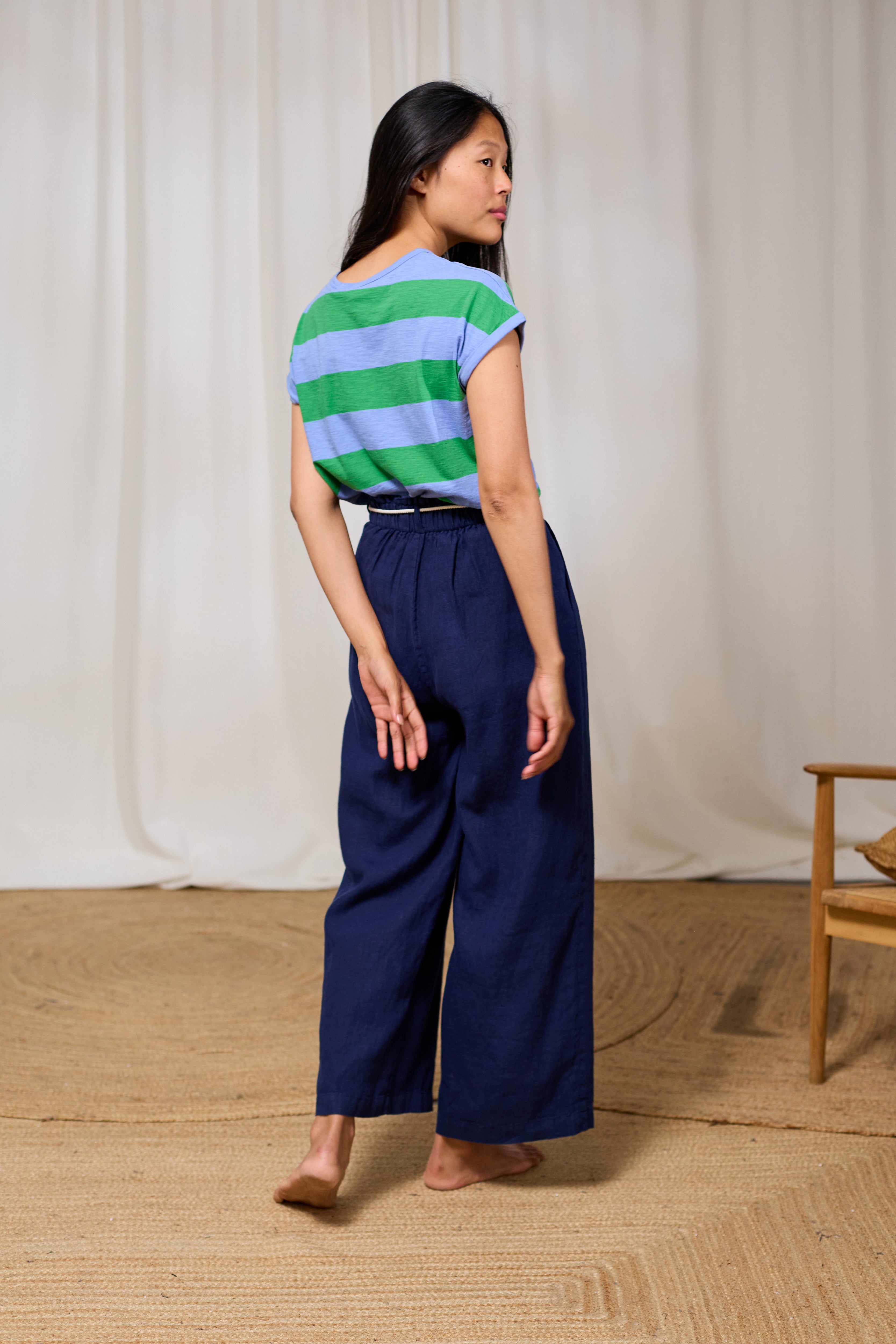 Woman wearing a colorful striped shirt and blue wide-leg pants standing against a white curtain.