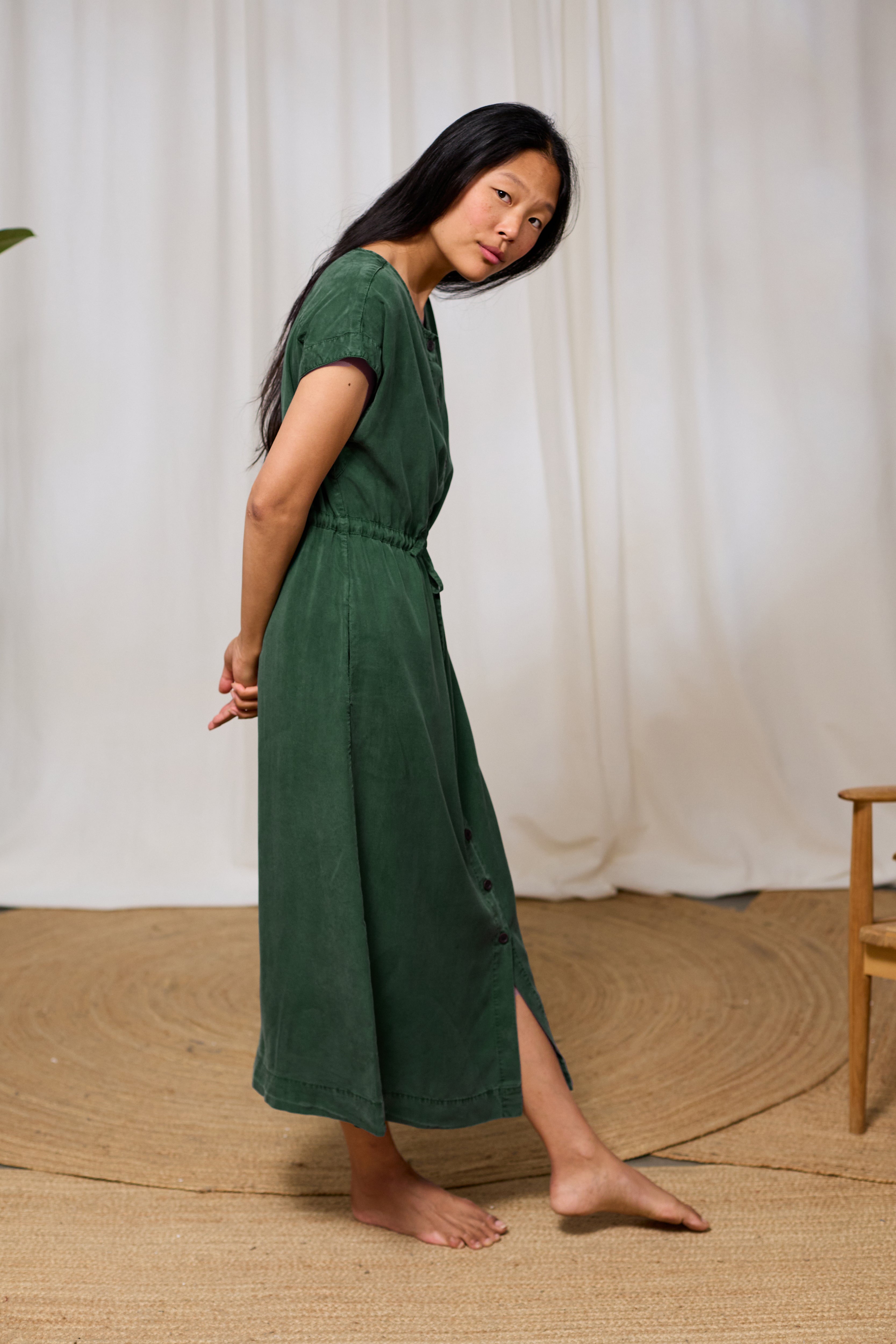 Woman wearing a green midi dress standing on a textured floor with a white curtain background