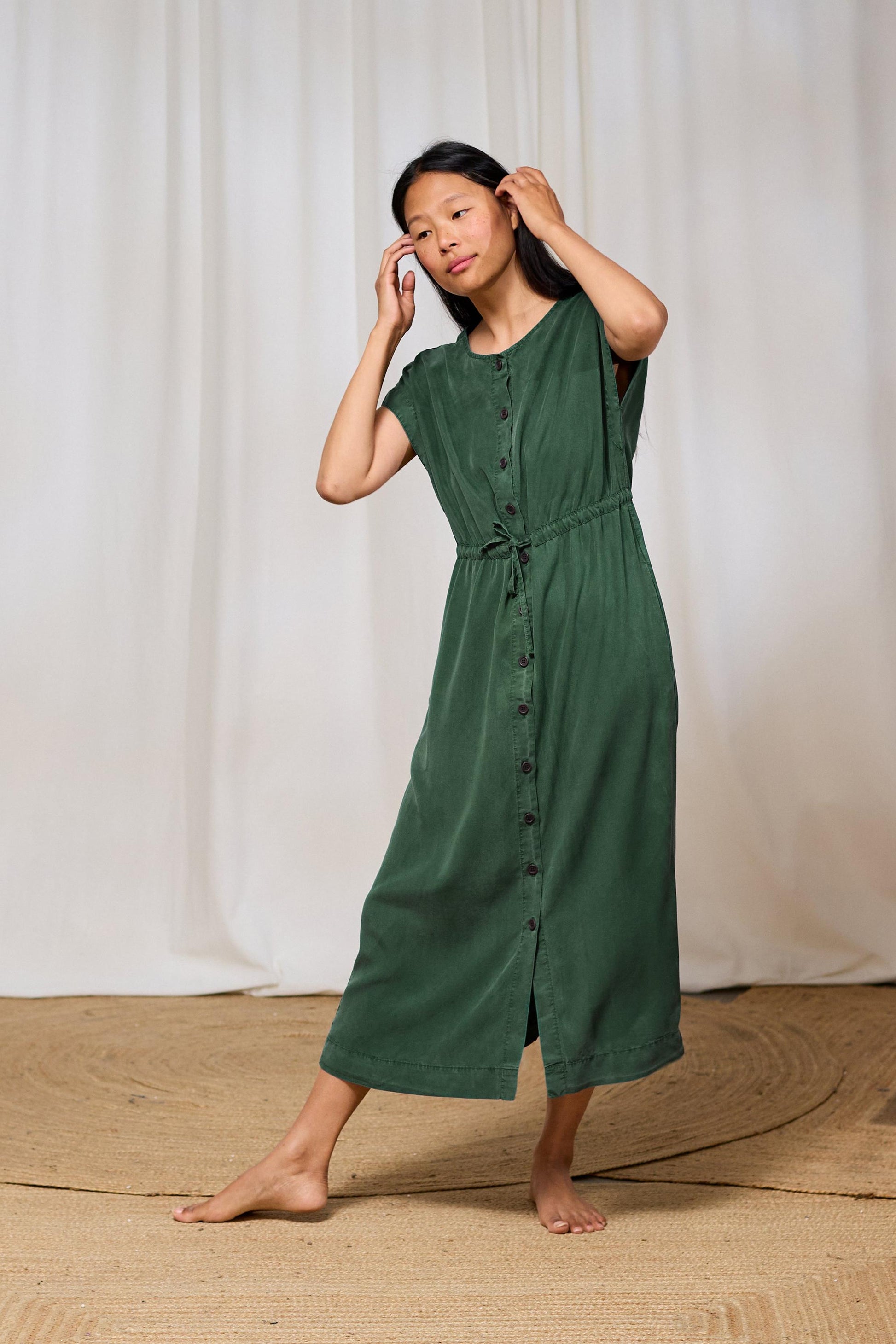 Woman wearing a green TWOTHIRDS dress standing against a white curtain background