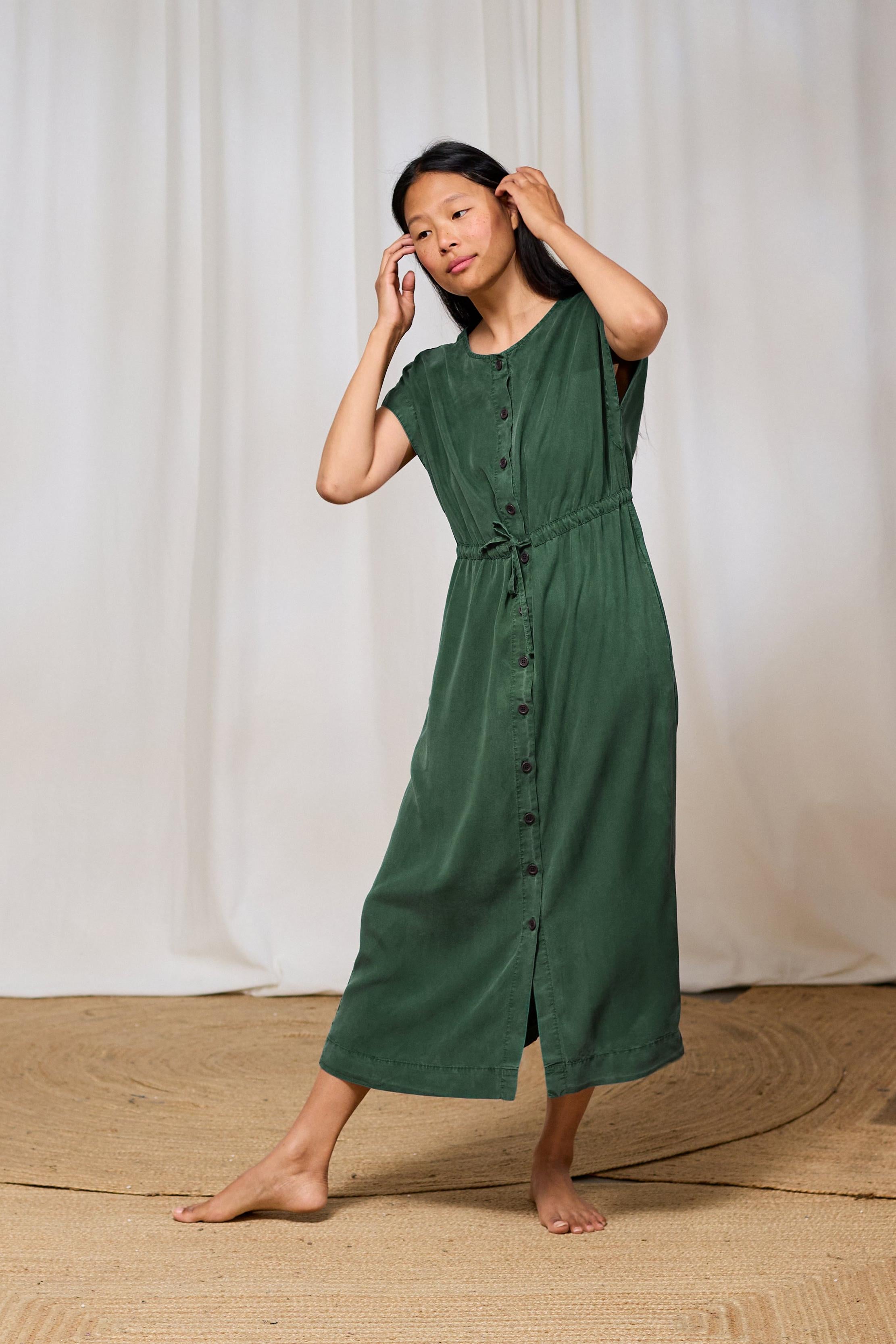Woman wearing a green TWOTHIRDS dress standing against a white curtain background