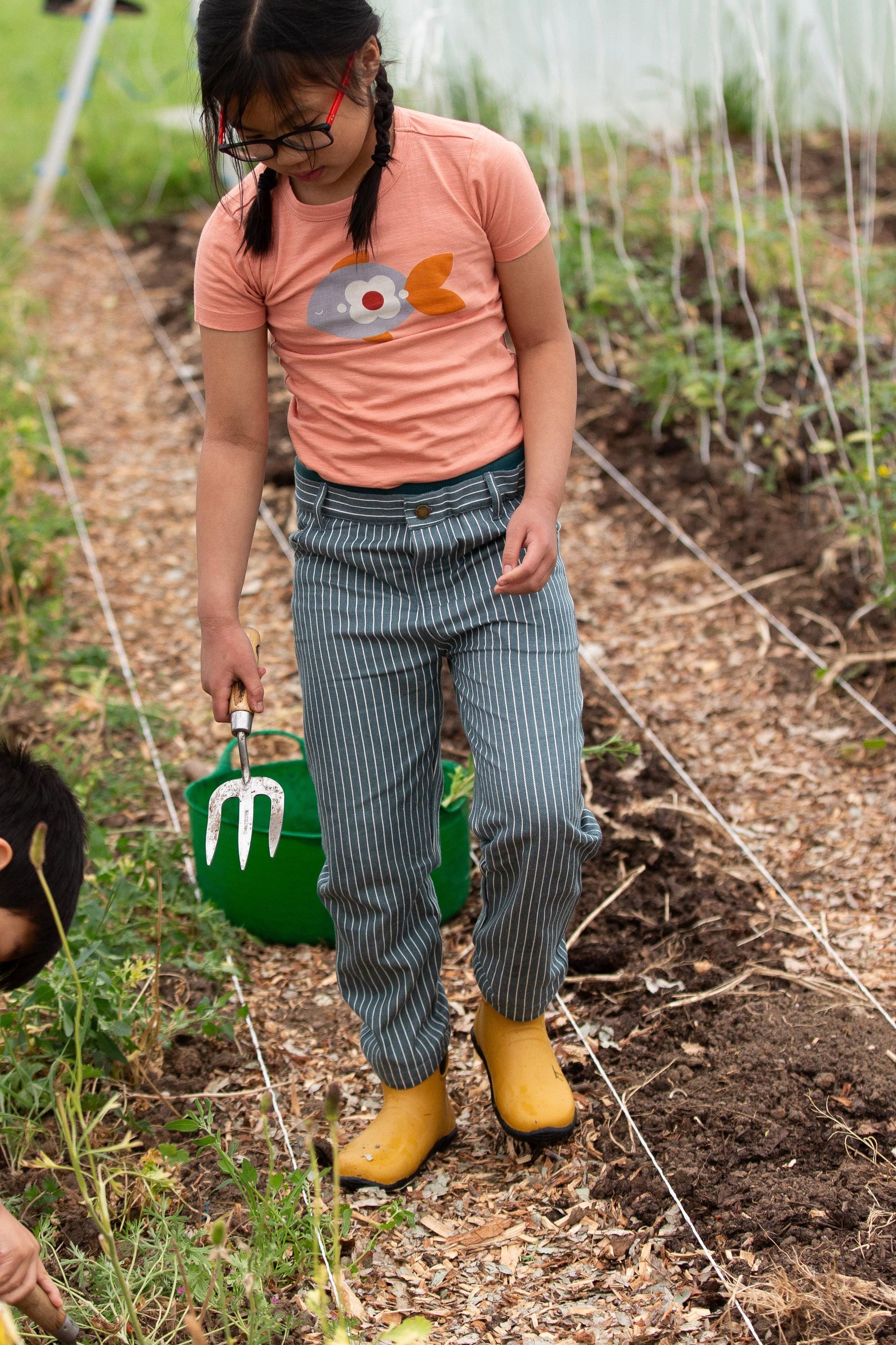 Child in a garden wearing a pink shirt with a cartoon design, striped organic cotton pants, and yellow boots.