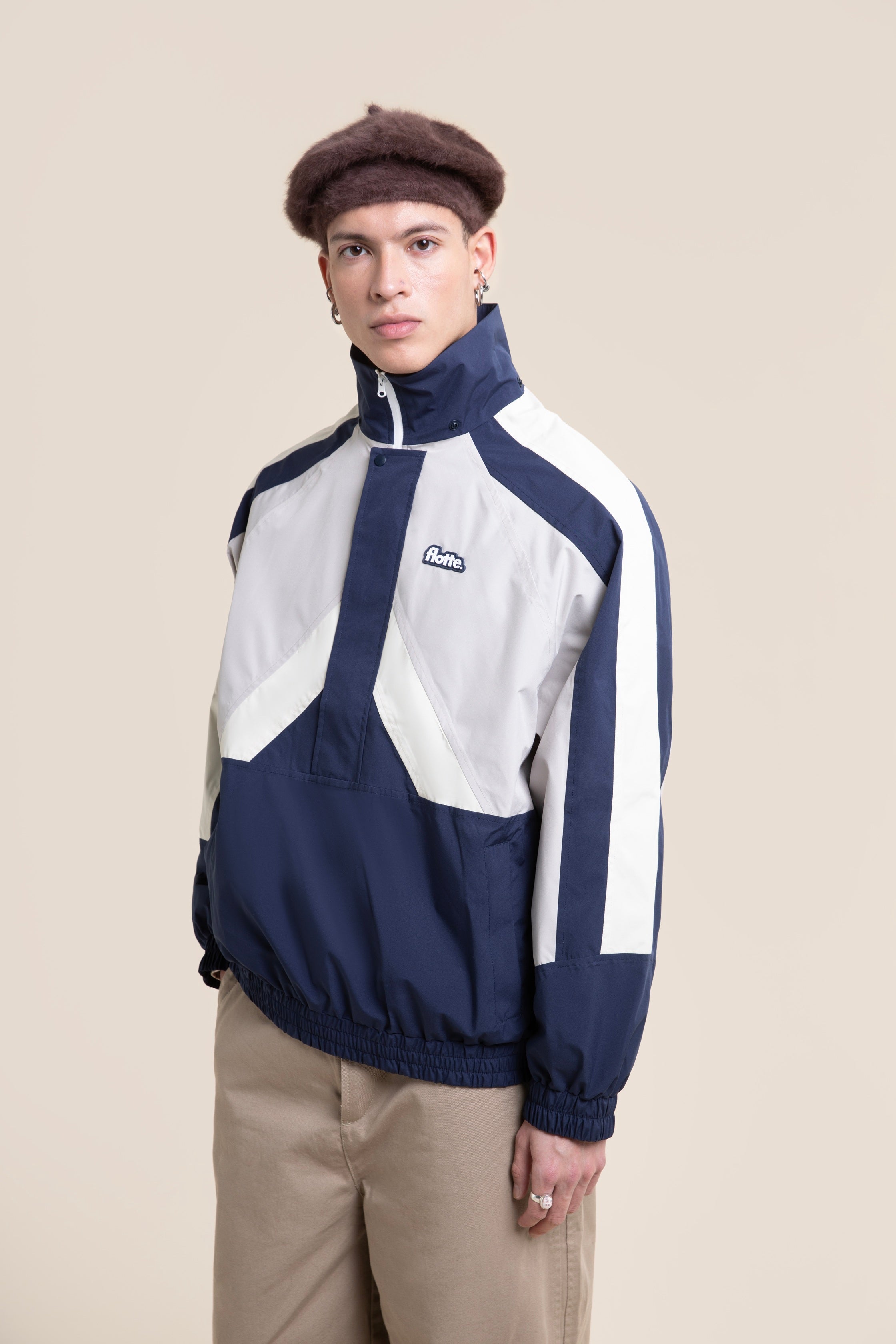 A person wearing a blue and white unisex waterproof jacket made from recycled polyester against a beige background.