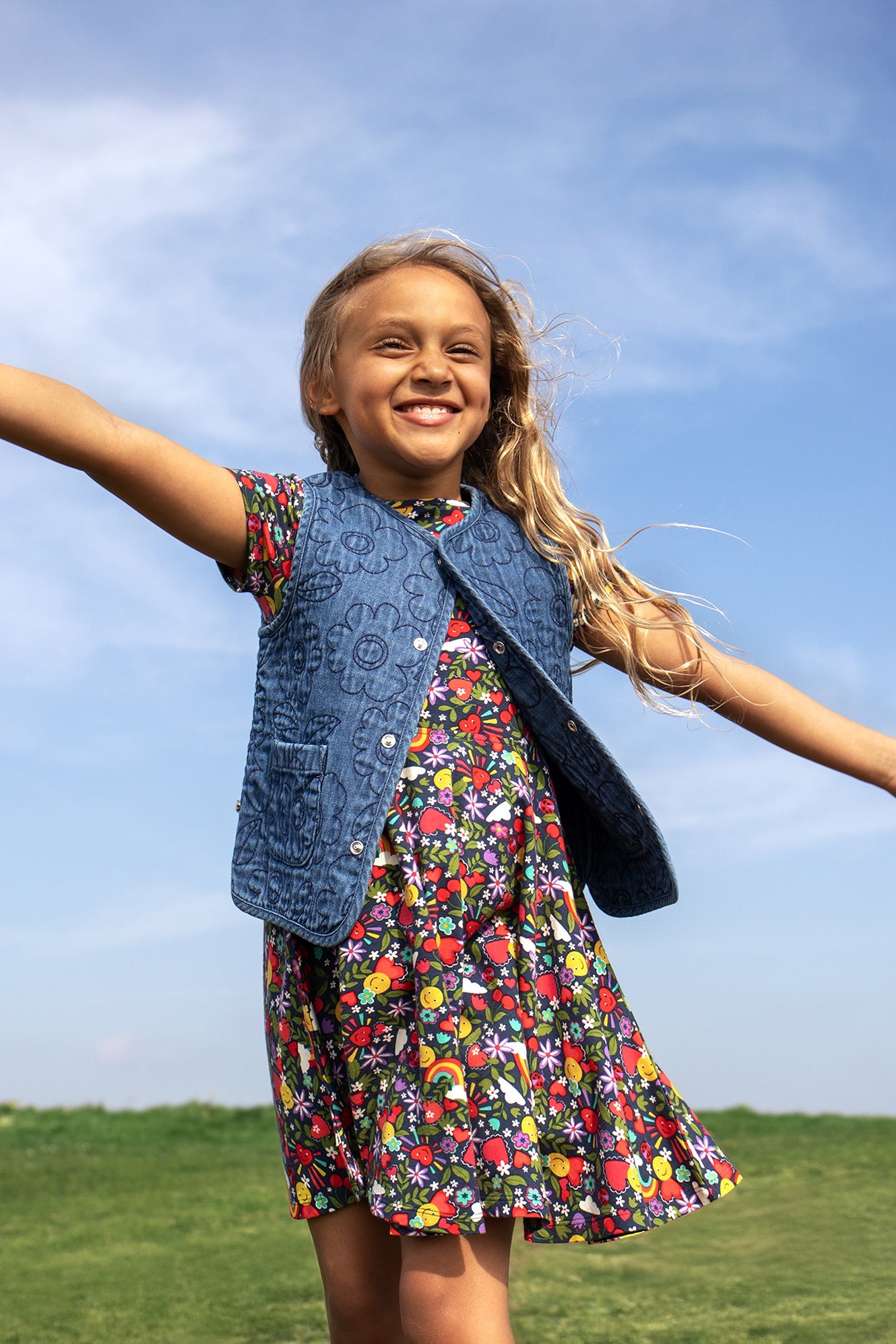 Young girl in a colorful Frugi dress with a blue vest standing outdoors against a clear blue sky.