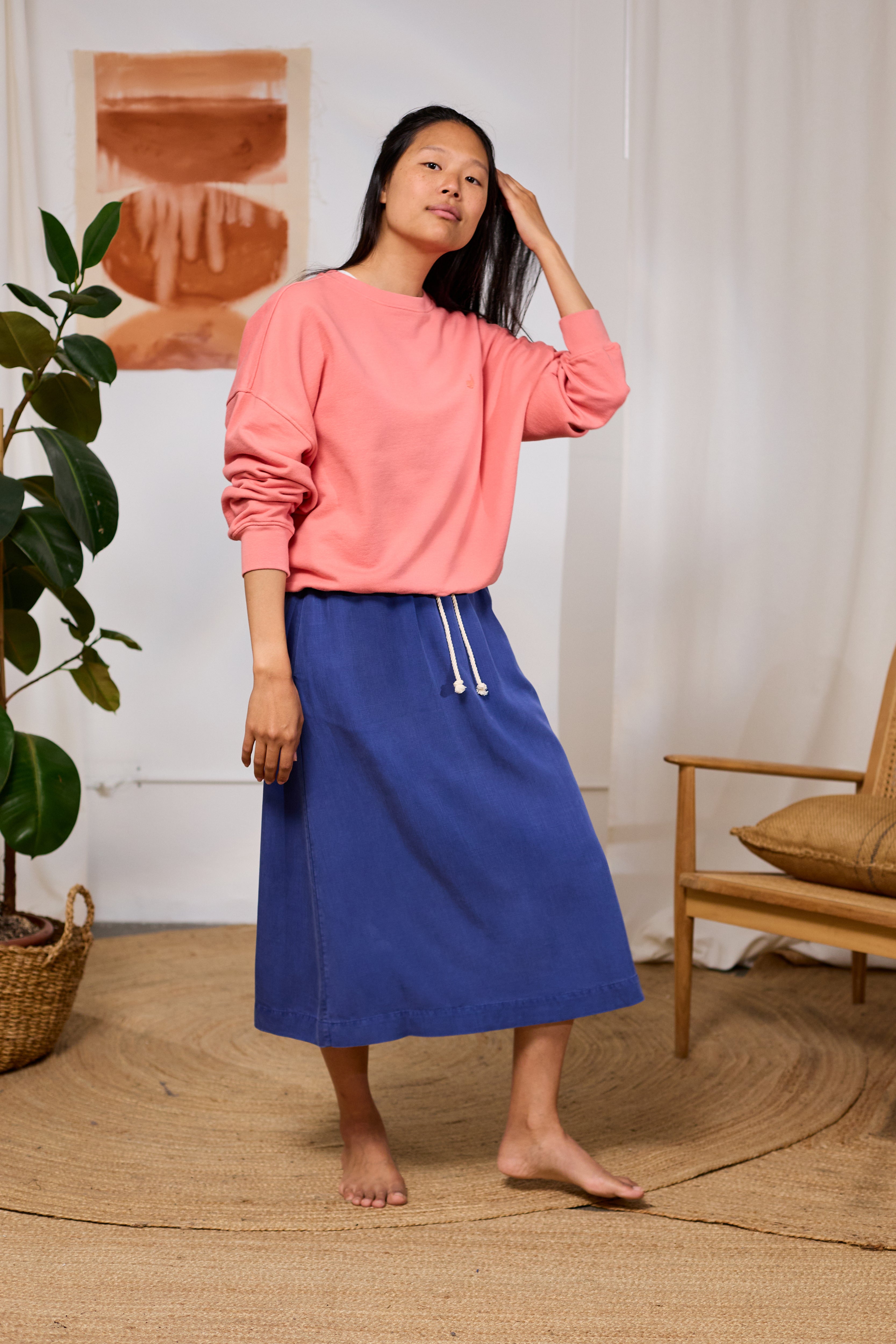 Woman wearing a pink organic TWOTHIRDS sweatshirt and blue skirt standing in a room with a plant and chair.
