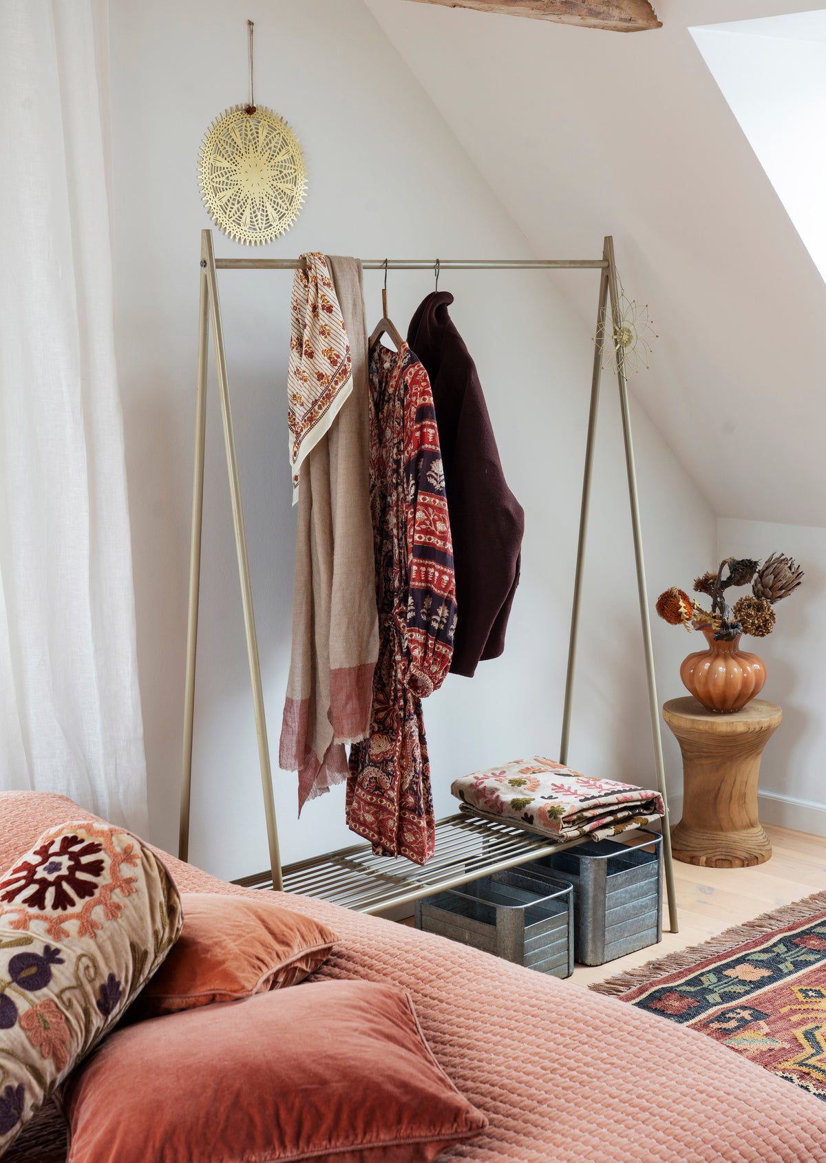 A brass A-frame clothing rack styled in a cozy bedroom corner with a slanted ceiling, holding patterned scarves and a dark jacket, with a folded matching textile on its lower shelf.