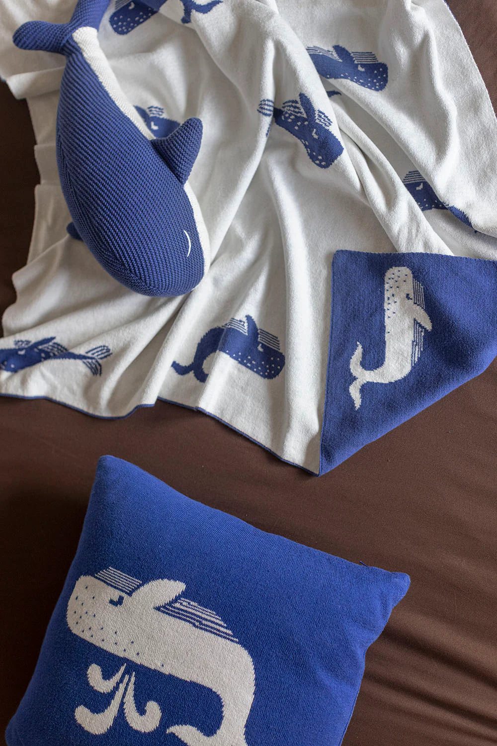 Knitted blue and white whale-patterned baby blanket and matching decorative pillow on a dark brown surface.