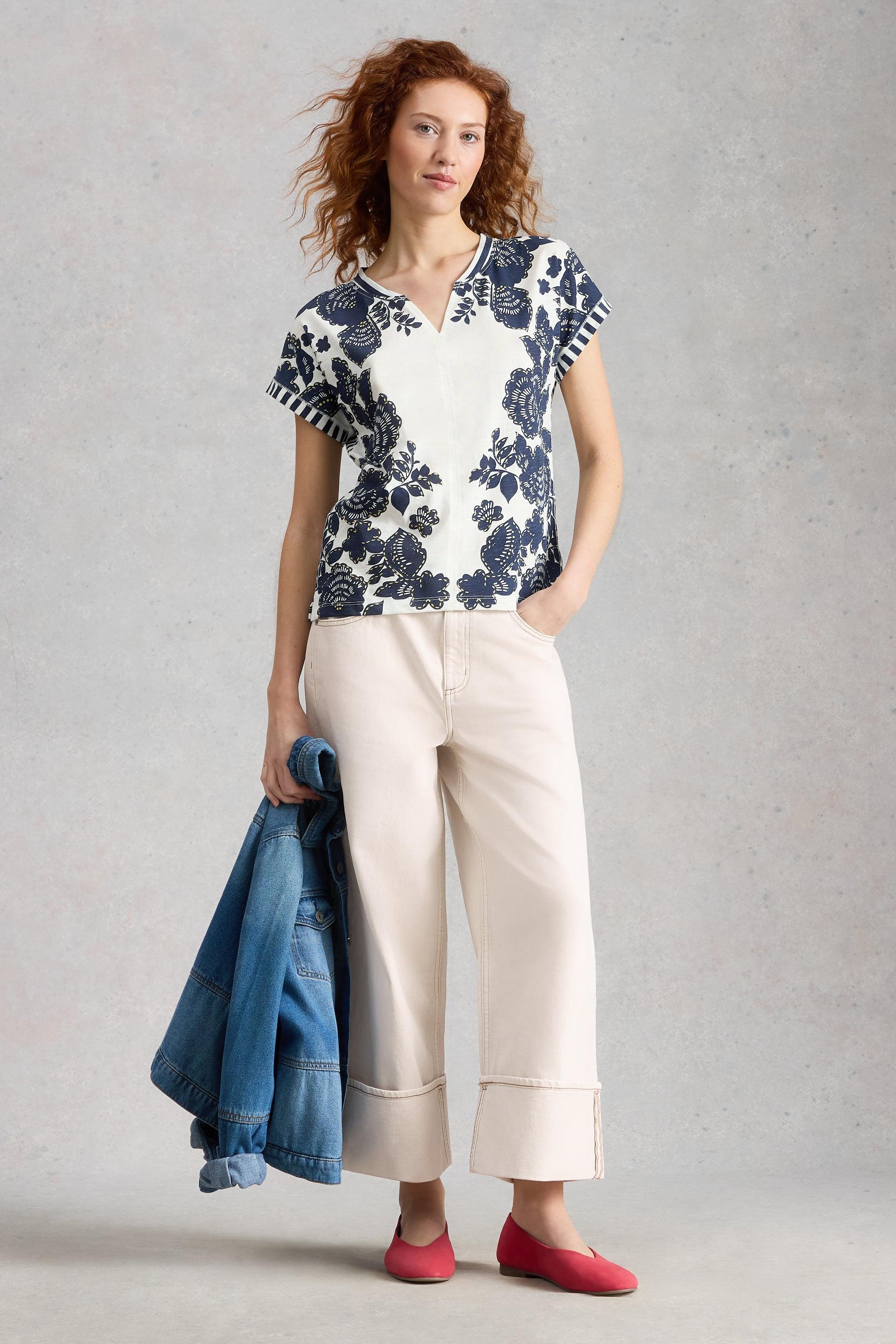 Woman wearing a floral White Stuff blouse and white pants, holding a denim jacket against a gray background