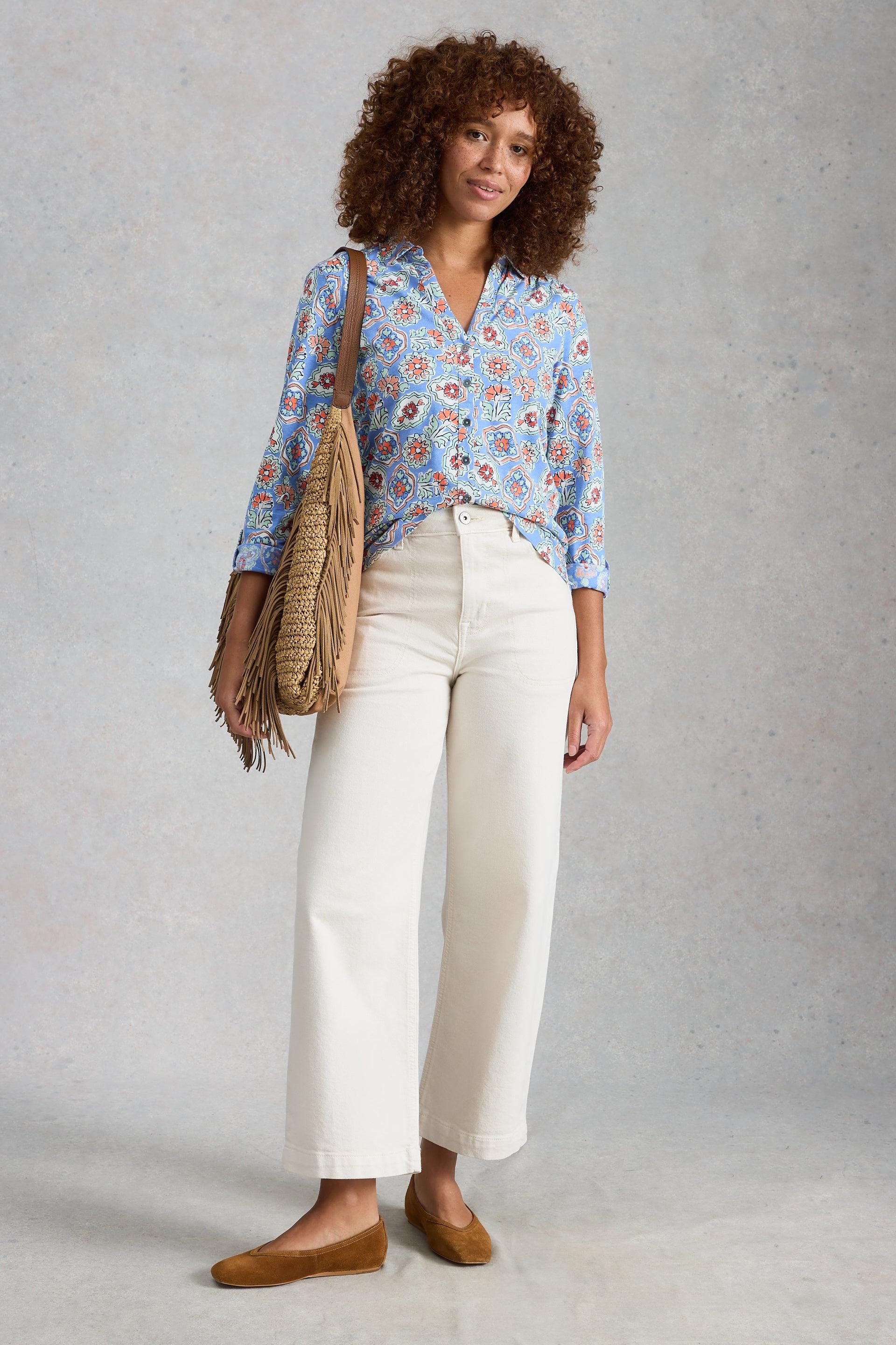 Woman wearing a blue floral blouse and white pants, holding a beige bag against a gray background