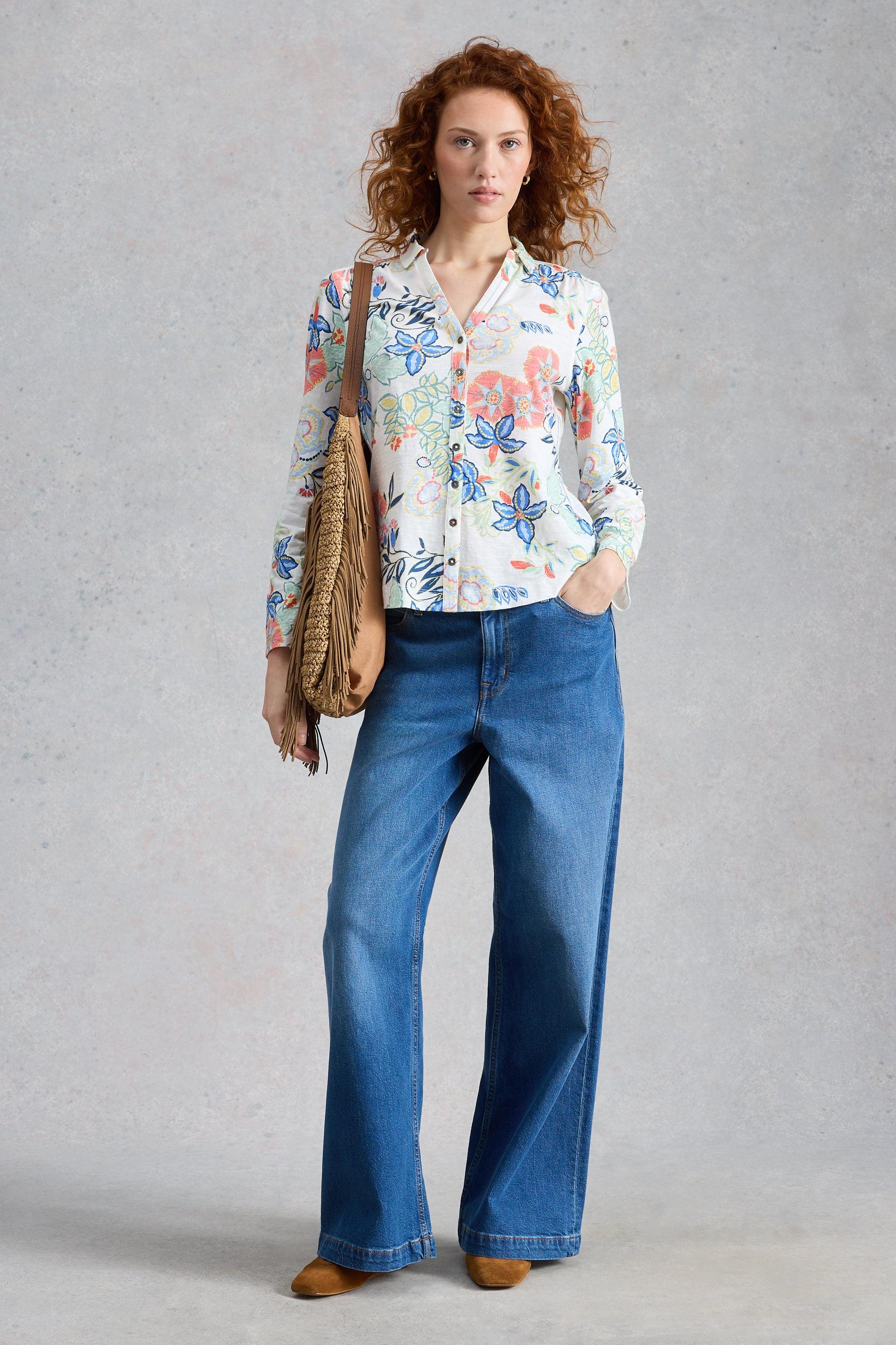 Woman wearing a floral White Stuff blouse and blue jeans, holding a brown bag against a gray background