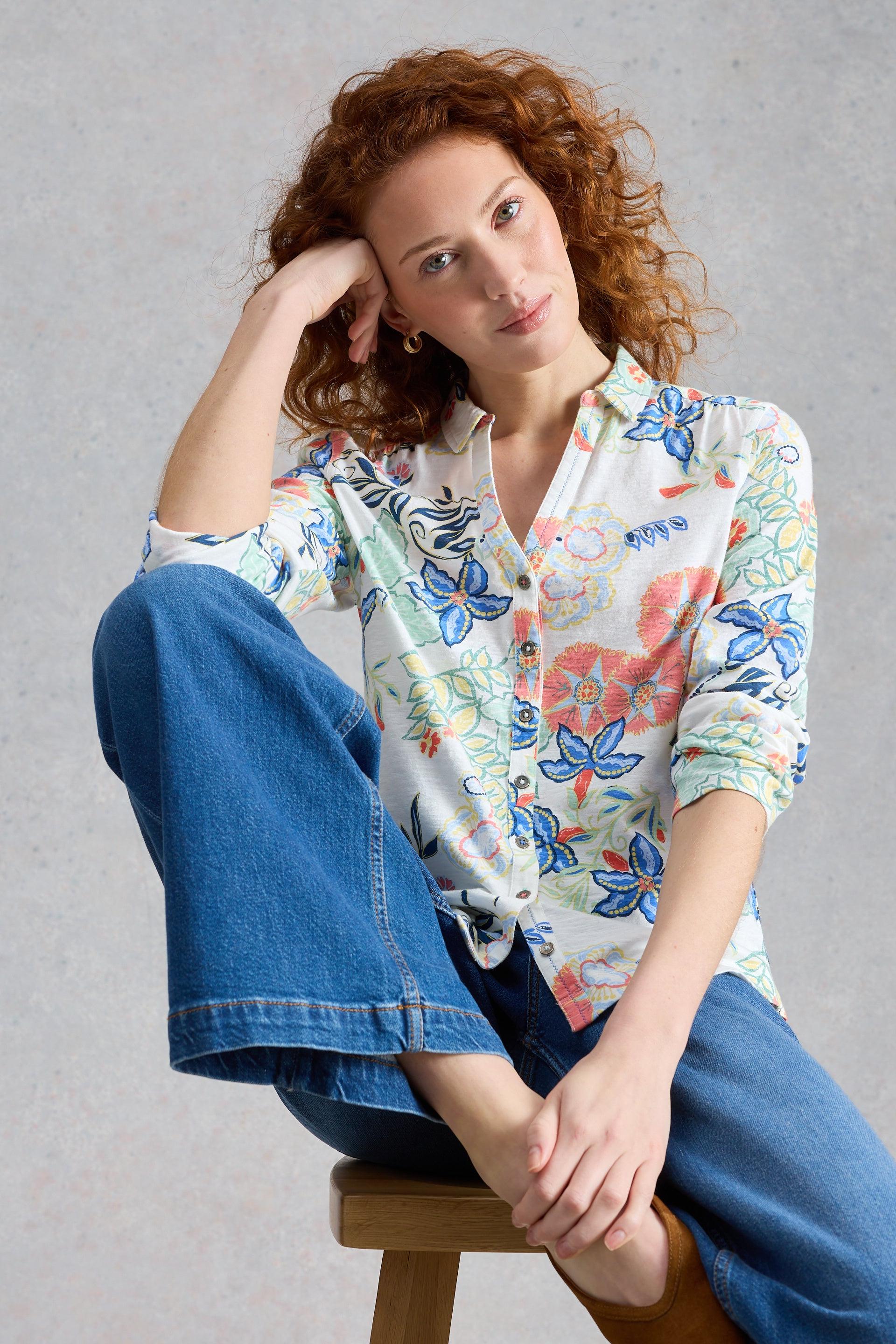Woman wearing a floral blouse and blue wide leg jeans sitting on a stool against a gray background