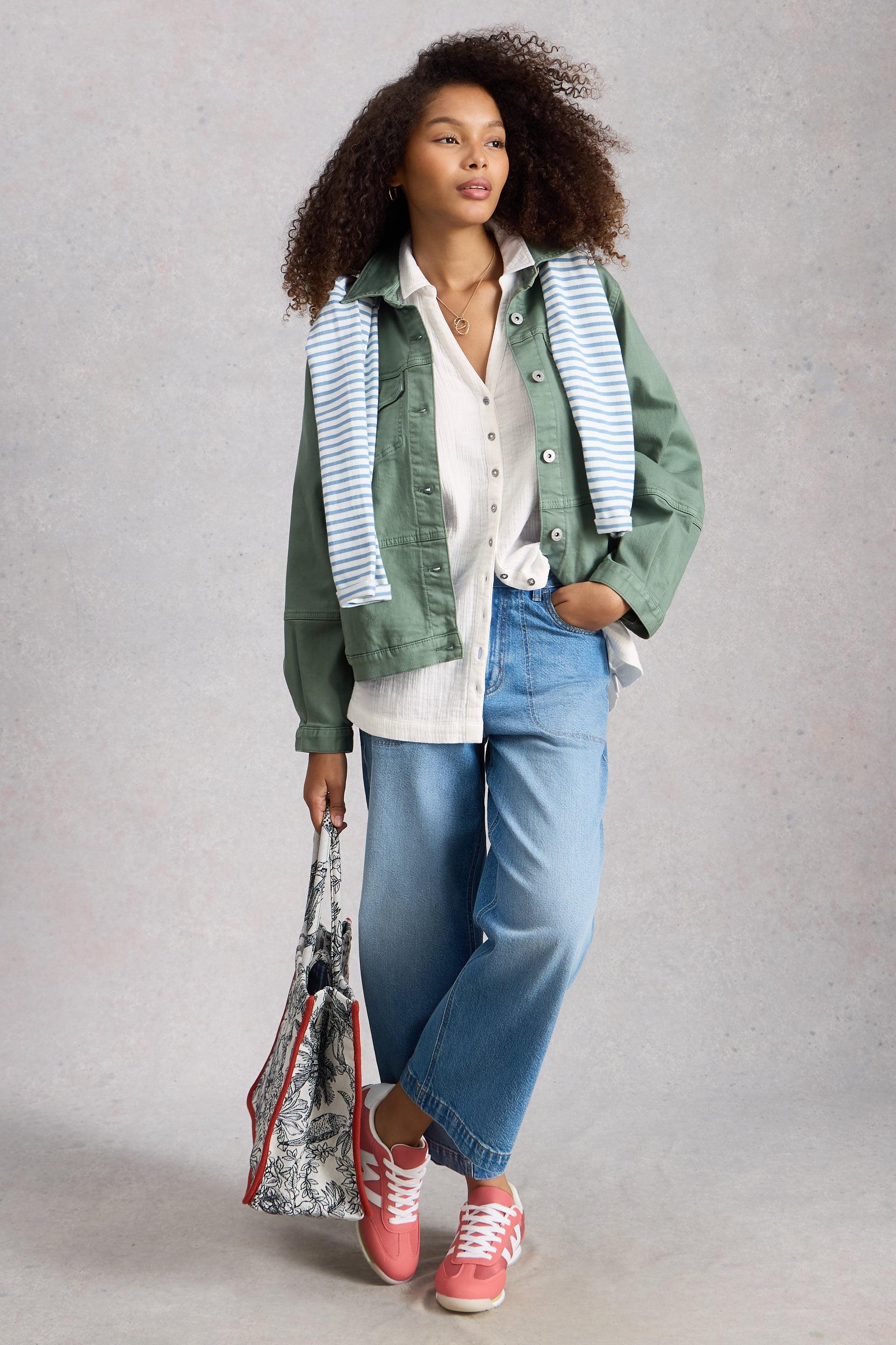 Woman wearing a green jacket, white shirt, blue jeans, and pink sneakers, holding a patterned bag against a gray background.
