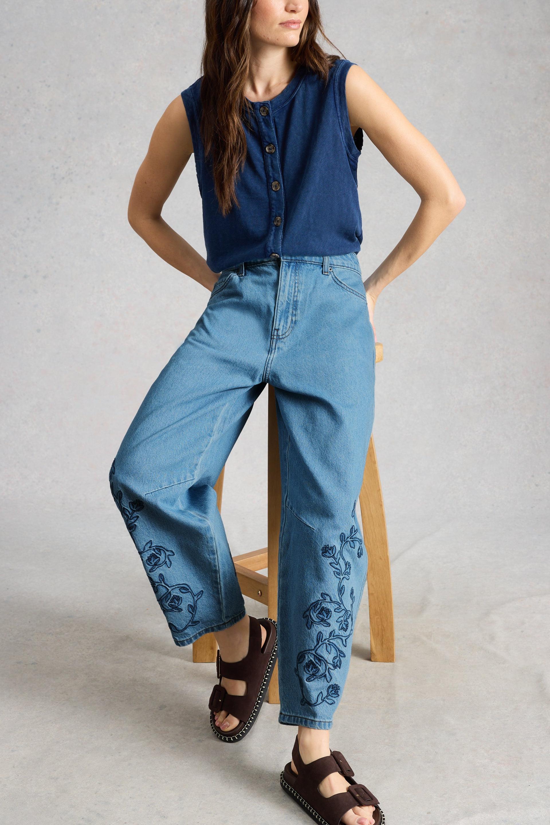 Woman wearing a blue sleeveless top and organic cotton jeans with floral embroidery on a gray background